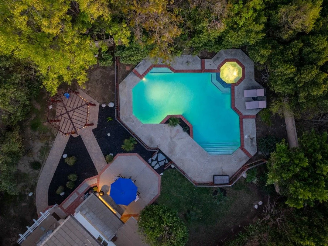 Aerial view of a backyard with a swimming pool, surrounding trees, a gazebo, a patio area, and outdoor furniture.