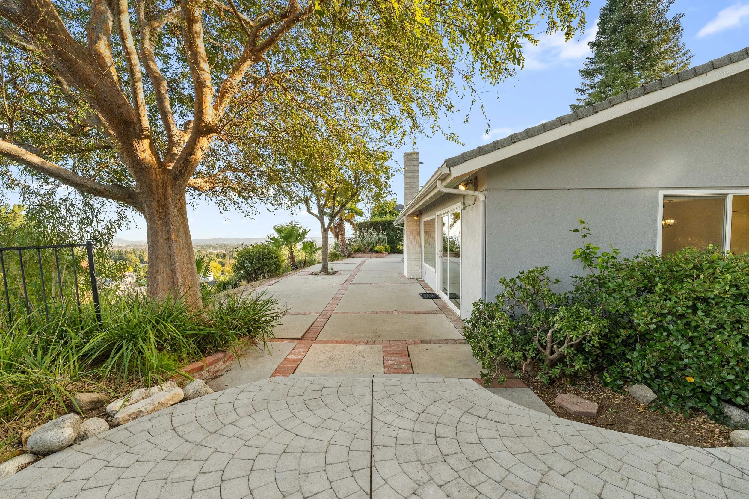 View of a backyard patio with stone walkway, bushes, trees, and a house with sliding glass door