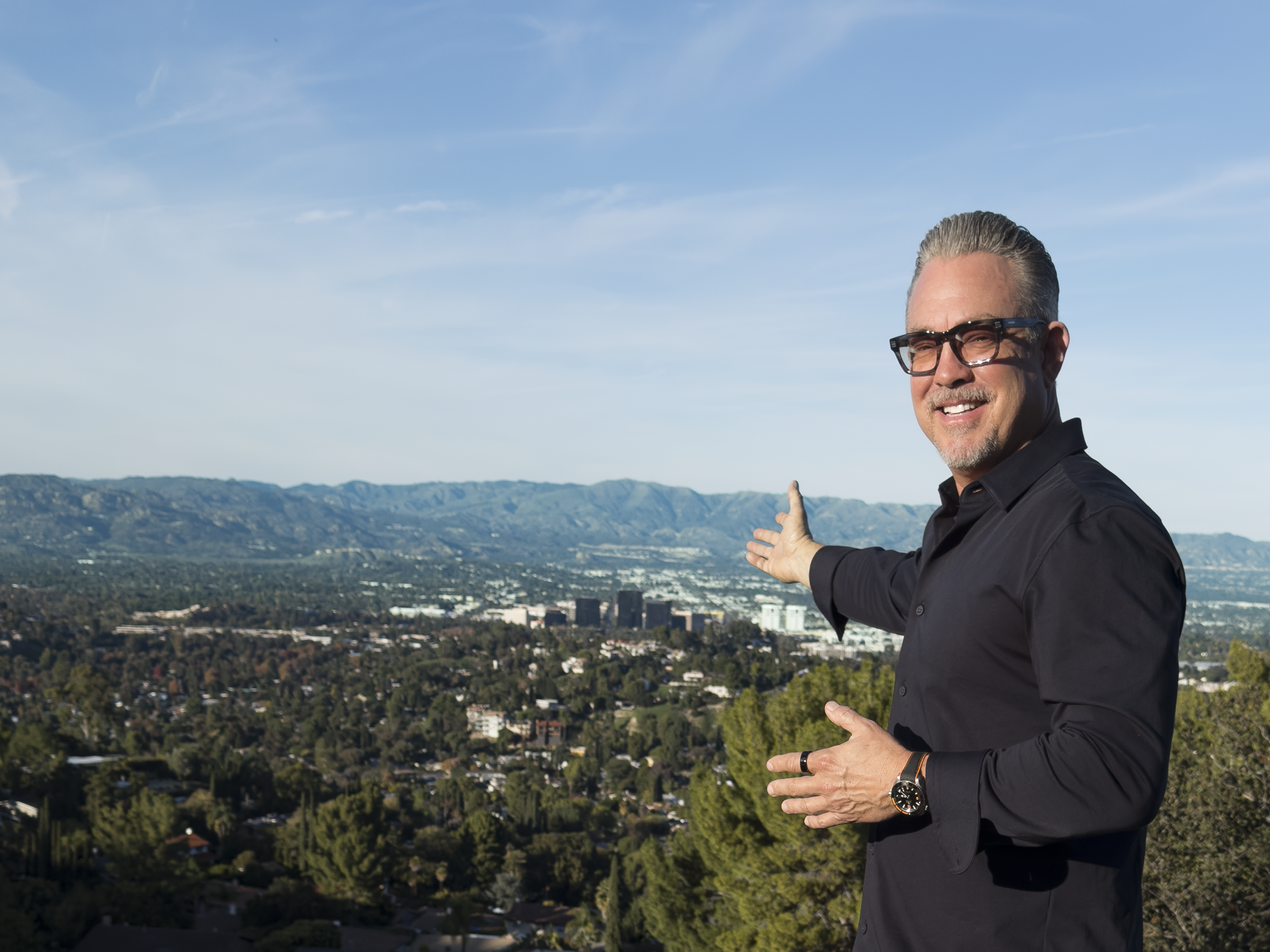 A man with gray hair, glasses, and a beard wearing a black shirt, standing outdoors on a hill, pointing toward a cityscape with mountains in the background.