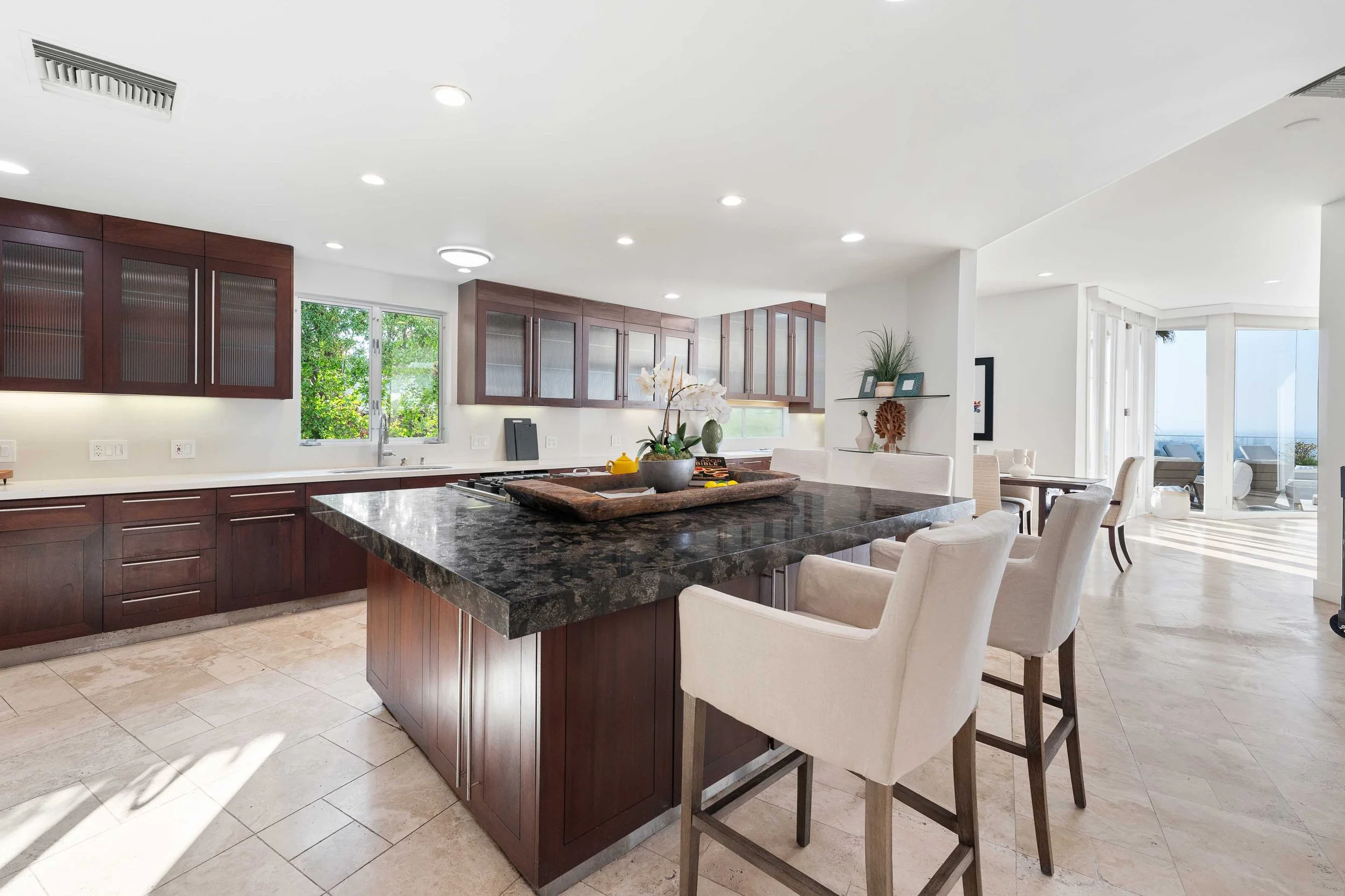 Bright, spacious modern kitchen with dark wood cabinets, large black granite island, beige tile flooring, and a window overlooking greenery. Adjacent dining area features white chairs and a glass table. Sunlit patio with outdoor furniture is visible 