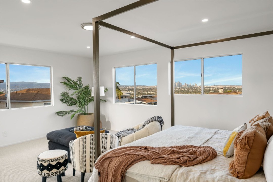 A bright bedroom with large windows showing a city view, a four-poster bed with tan and white bedding, a black ottoman, a white and black patterned stool, a beige armchair, a large potted plant, and a small lamp.