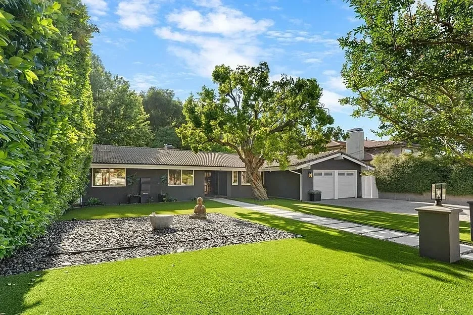 A well-maintained backyard with green grass, a gravel area with a decorative statue, a large tree, and a house with a gray exterior and white garage doors in the background.