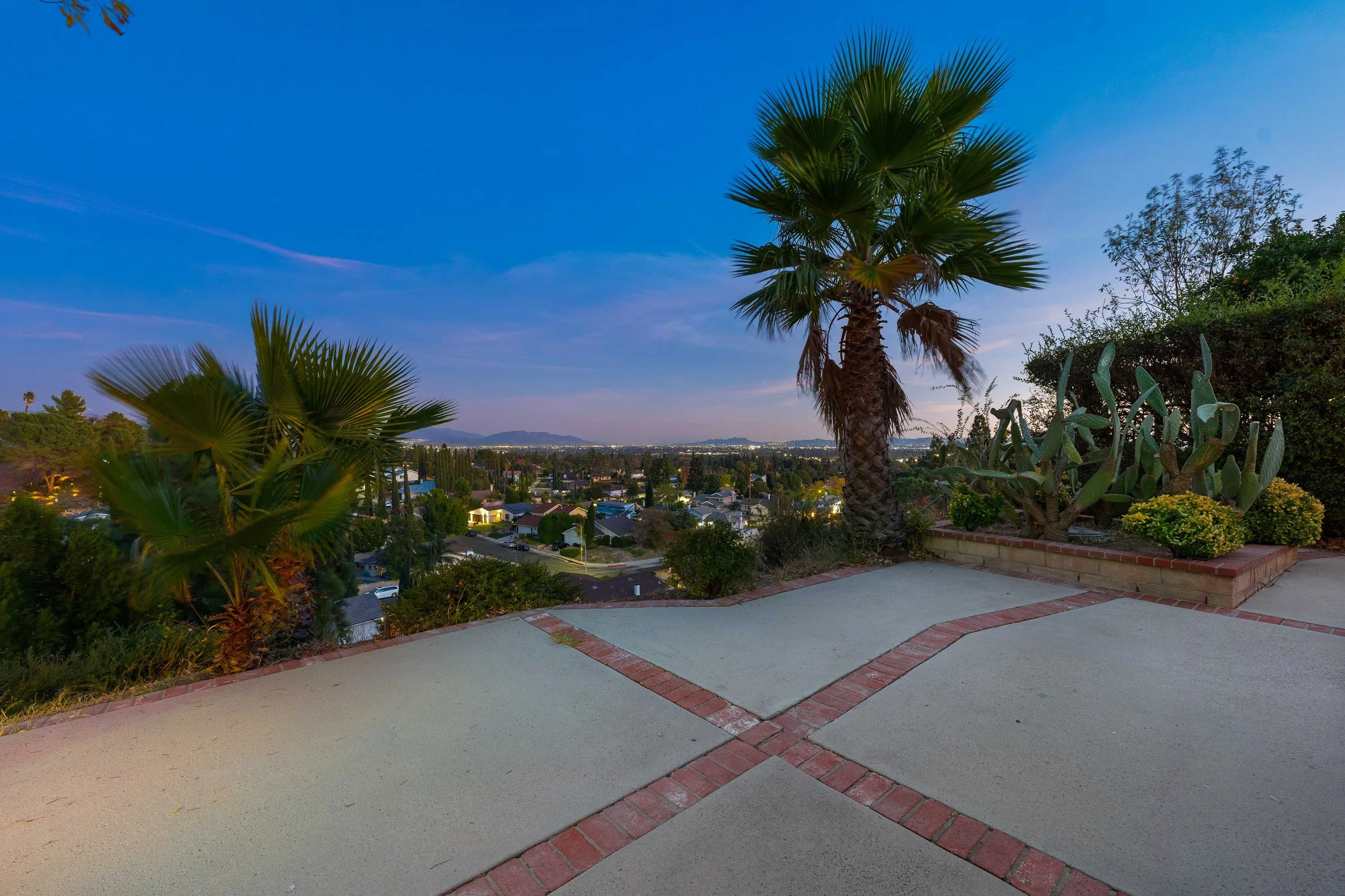 A view of a suburban neighborhood with palm trees, desert plants, and city lights in the distance during dusk or dawn, with a paved patio in the foreground.