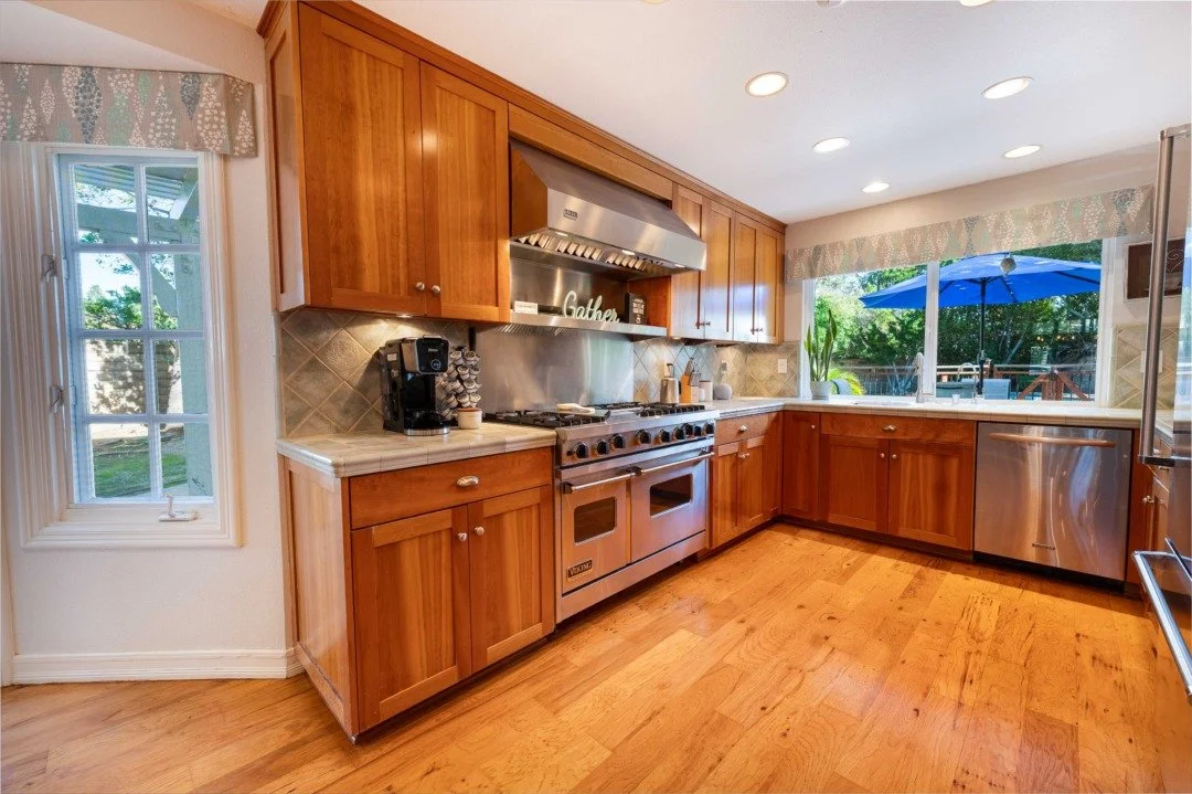 Kitchen with wooden cabinets, stainless steel stove and dishwasher, coffee maker, and large window with blue umbrella outside.