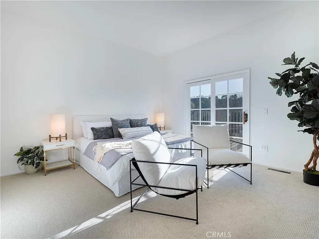 Bright bedroom with white walls, a bed with white and gray bedding, two modern side tables with lamps, a potted plant, a large window, two white chairs with black metal frames, and a large potted plant in the corner.