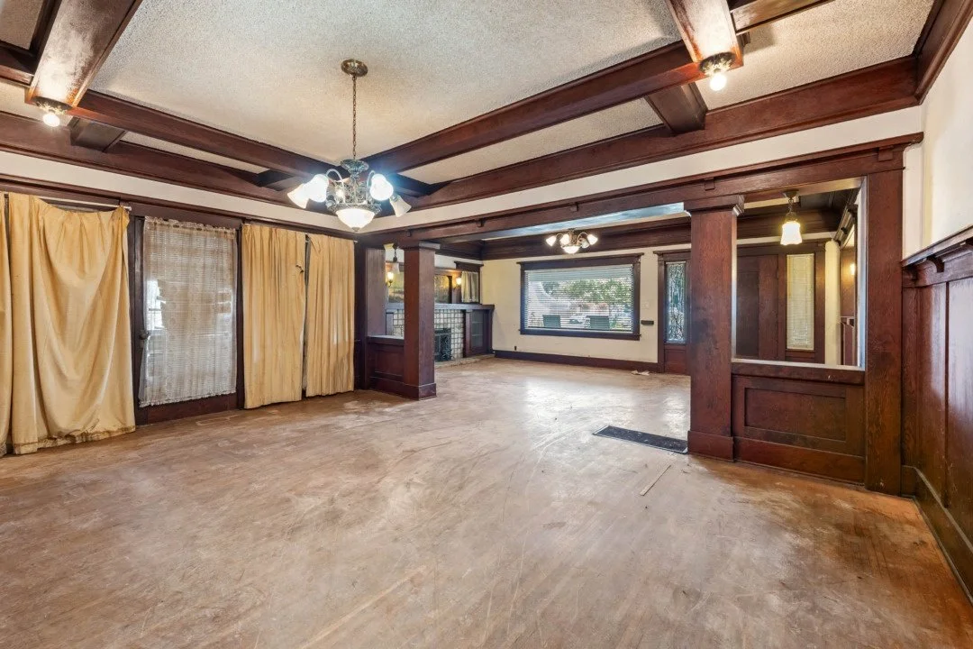 Empty living room with hardwood floors, large window, dark wood trim and paneling, and draped curtains.