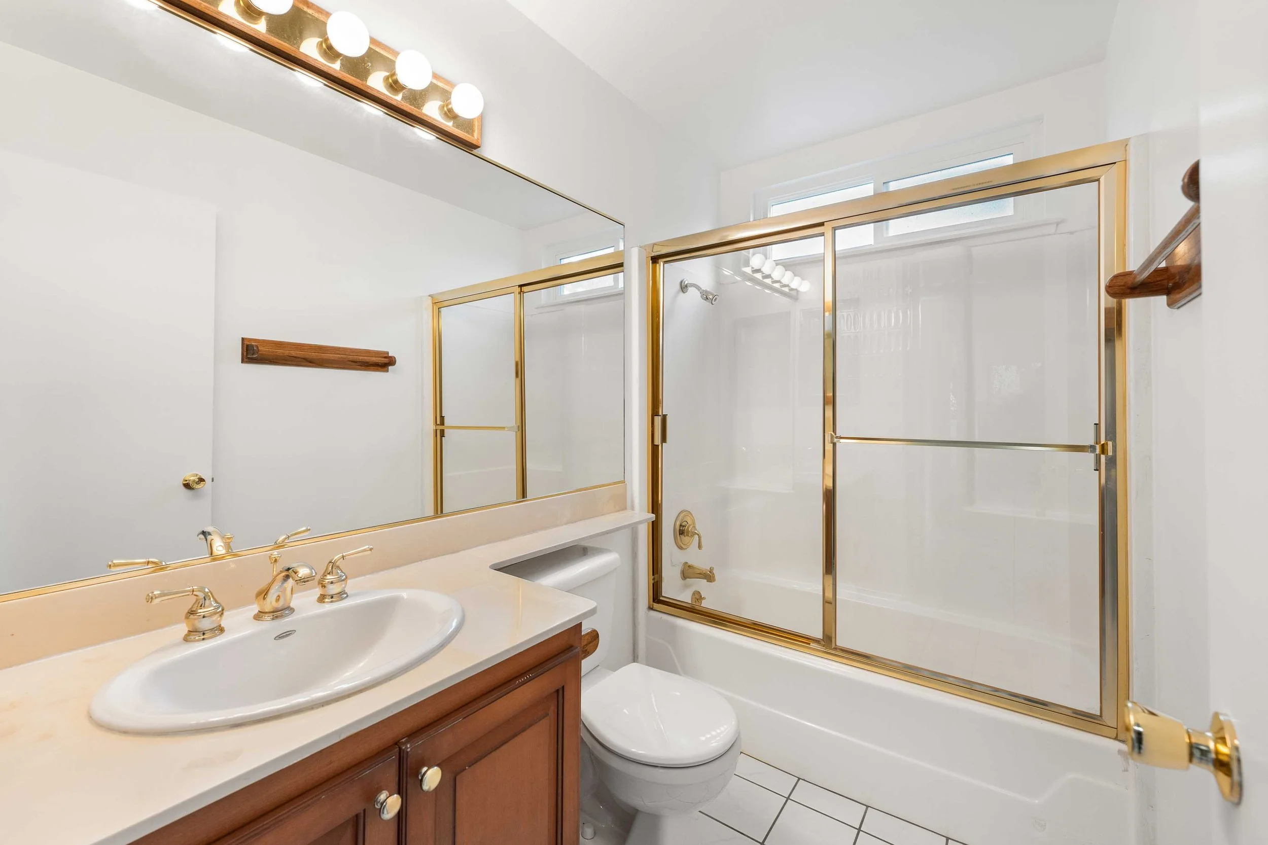 Bathroom with a white sink, wooden vanity, toilet, glass shower with gold frame, large mirror, and a small window