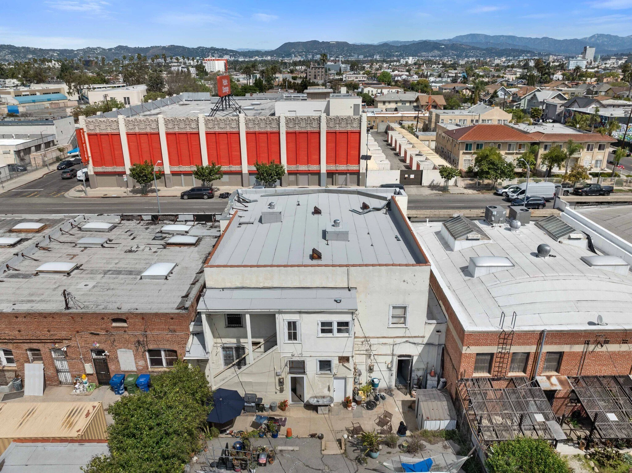 Aerial view of a city neighborhood showing rooftops of buildings, streets, cars, and a large shopping mall with a red facade in the background, mountain range in the distance.