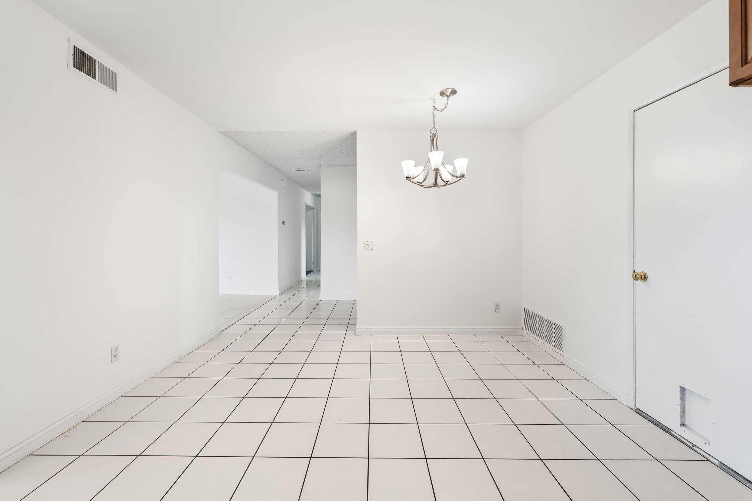 Empty dining area with white tiled floor, white walls, a chandelier, and a door on the right wall.