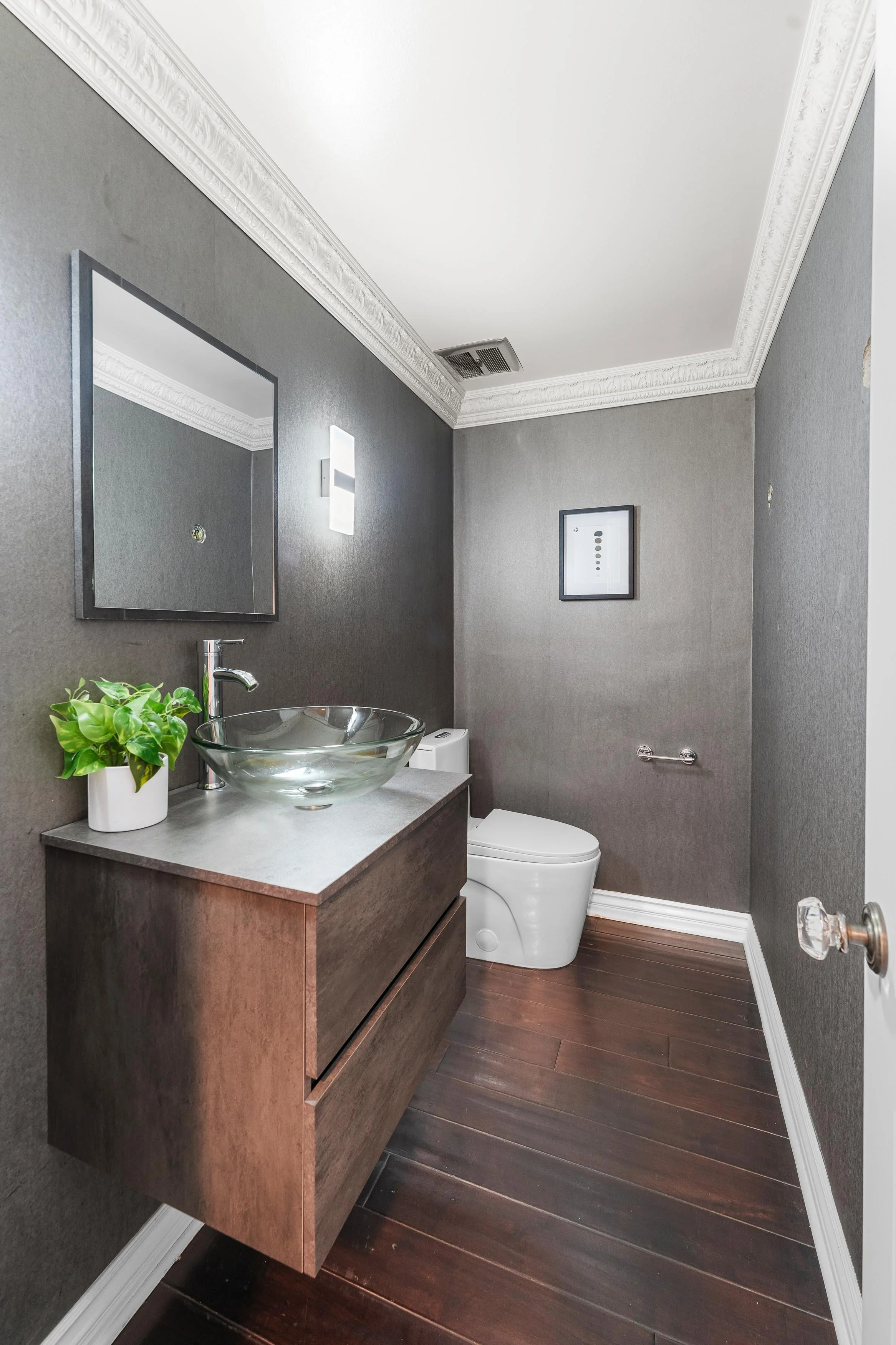 Small modern bathroom with dark gray walls, white crown molding, a floating wooden vanity with a glass vessel sink, a mirror, a potted plant, a white toilet, a framed picture, and wood flooring.