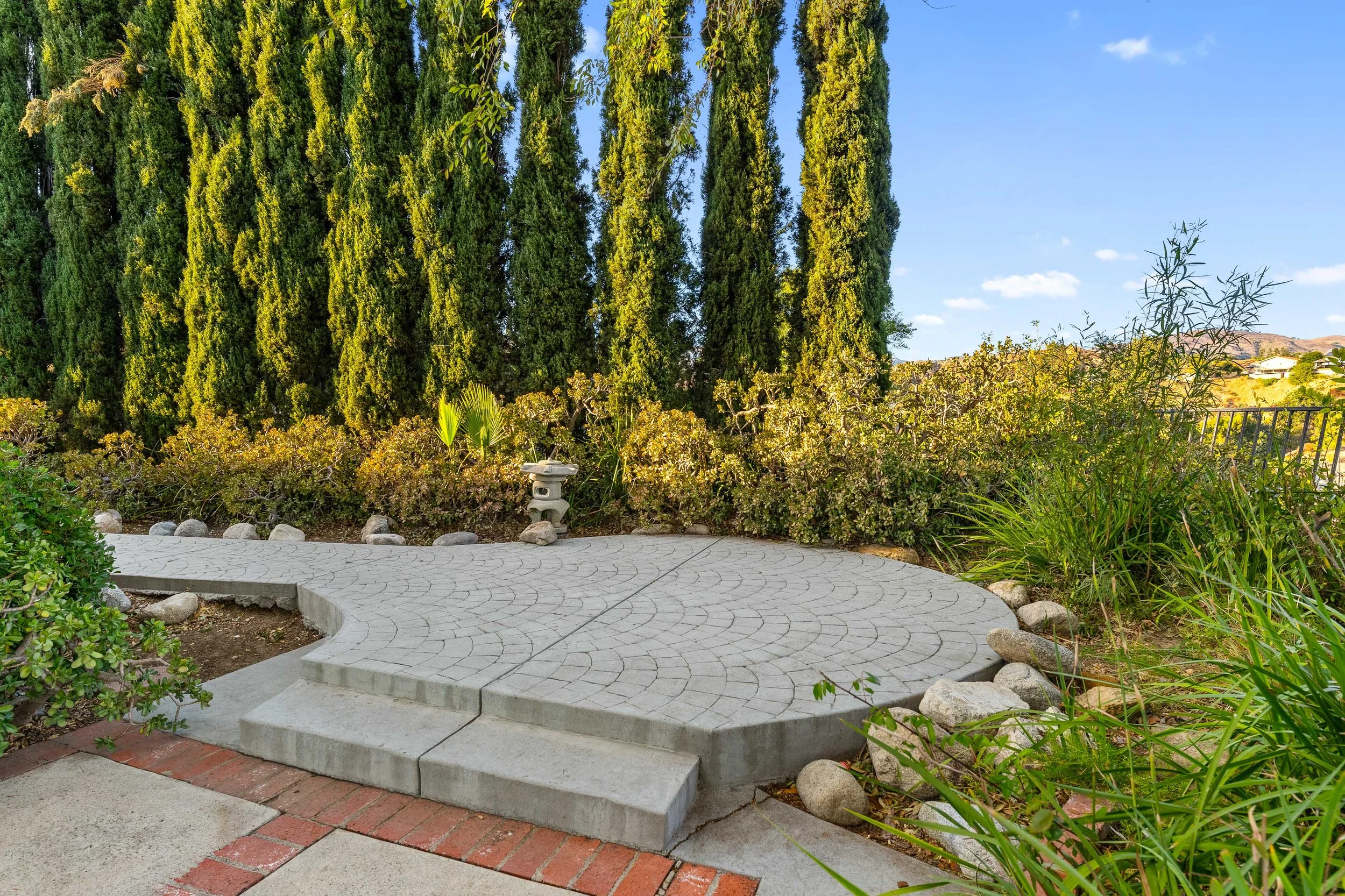 A small, circular concrete patio with steps leading up to it, surrounded by green plants, bushes, and tall trees, under a blue sky with a few clouds.