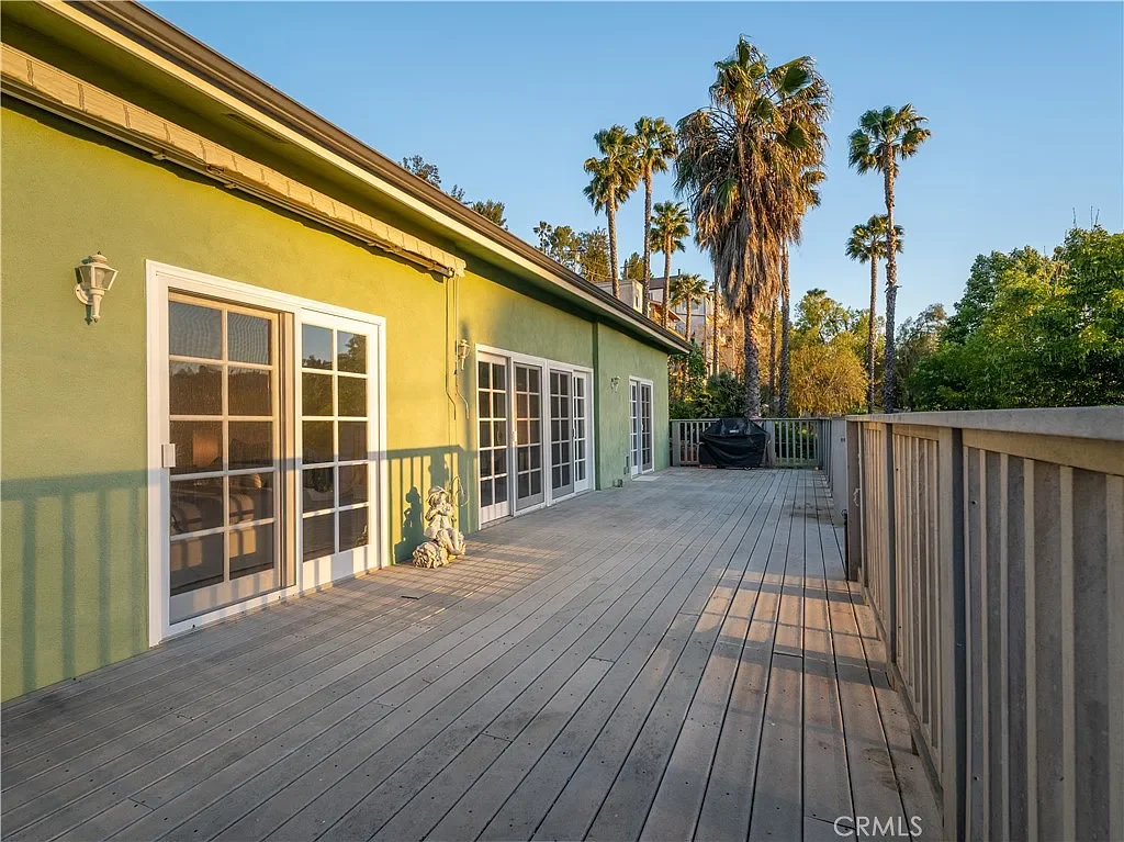 A spacious outdoor wooden deck attached to a green house with white-framed sliding glass doors, surrounded by palm trees and lush greenery, during late afternoon sunlight.