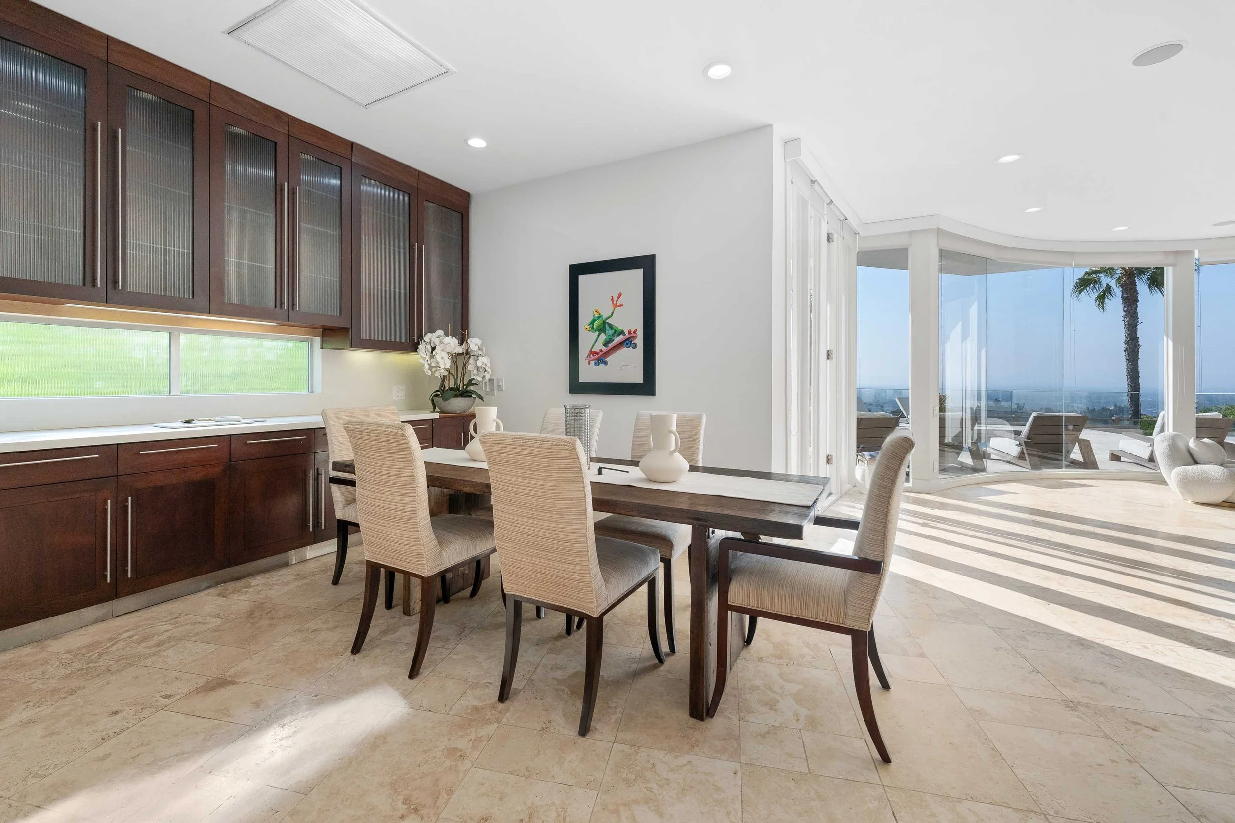 Dining area with a rectangular wooden table surrounded by six upholstered chairs, a dark wood cabinet with glass-paneled upper doors, a large window behind the cabinet, a framed colorful art piece on the white wall, a white flower arrangement, and a 