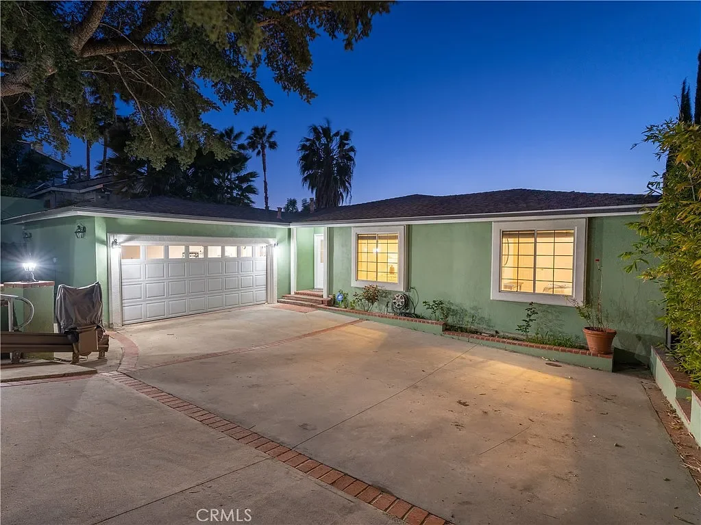 Nighttime view of a house's driveway with a closed garage door, illuminated by exterior lights. The house has green walls, yellow-lit windows, and a small garden bed with potted plants and garden decorations.