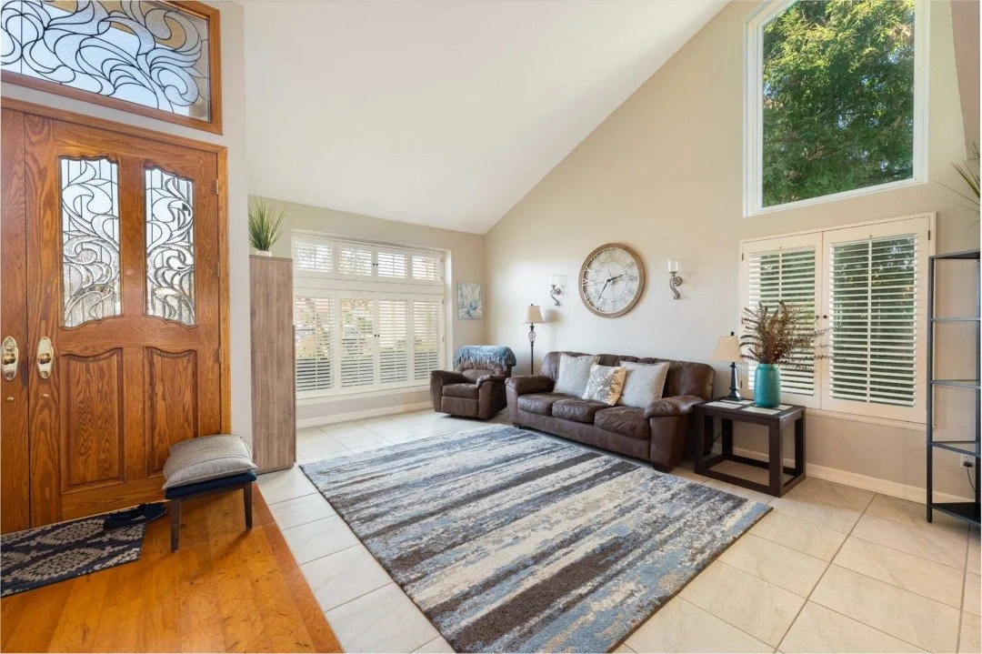 Living room with a brown leather sofa, white wall clock, side table with a lamp and vase, window with shutters, and a patterned area rug.