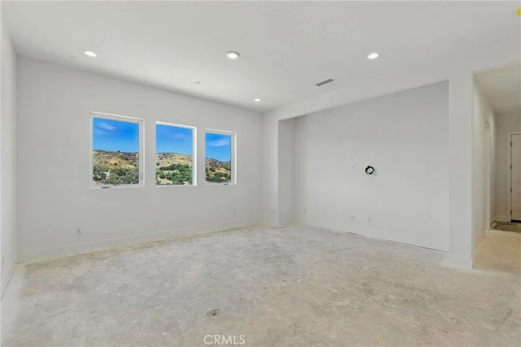 Empty room with white walls, three windows showing a view of hills and blue sky, and recessed ceiling lights.