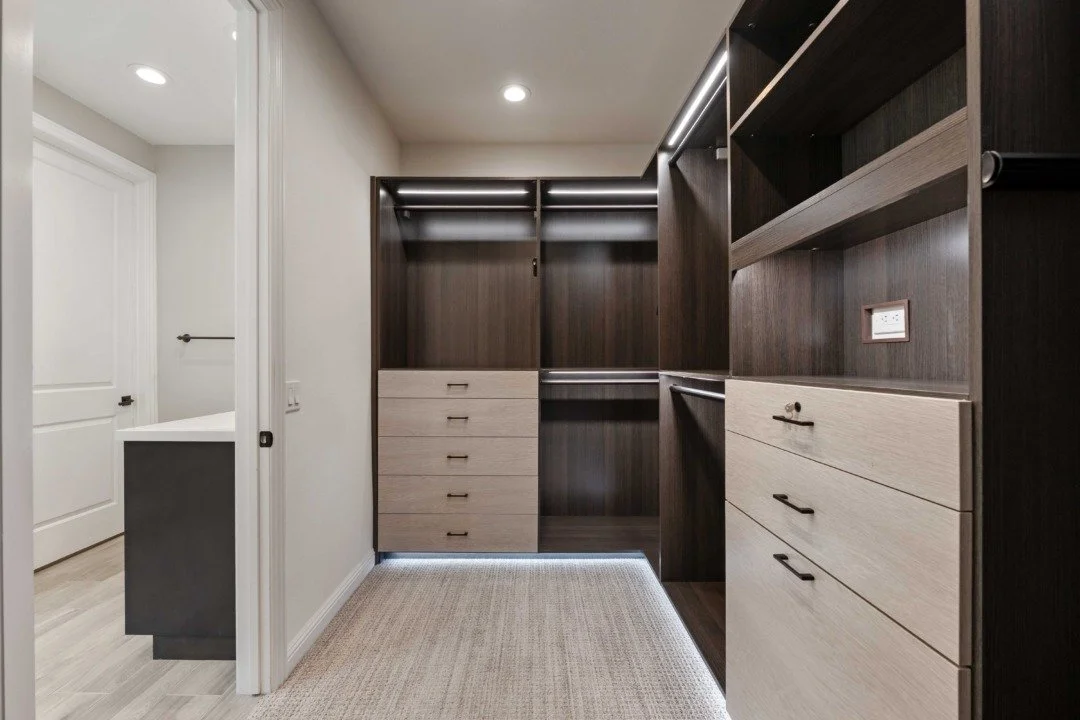 Empty walk-in closet with dark wood shelving and drawers, illuminated by recessed lighting, with a beige carpet and a doorway leading to a white-bathroom area.