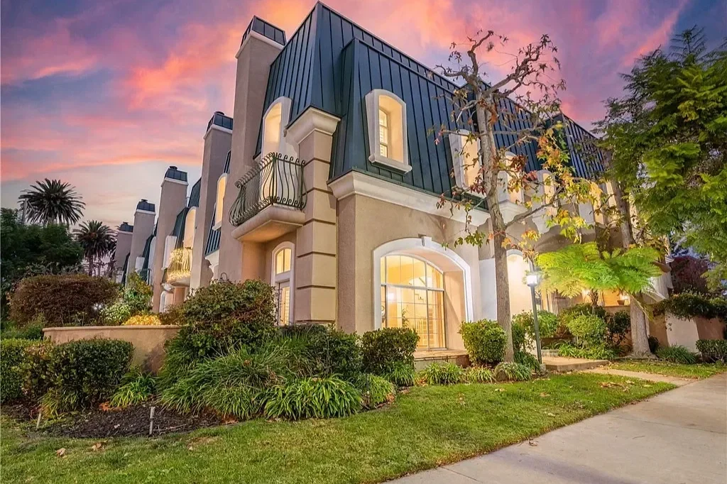 A row of elegant townhouses with large arched windows and small balconies, surrounded by landscaped greenery and trees, during sunset with a colorful sky.