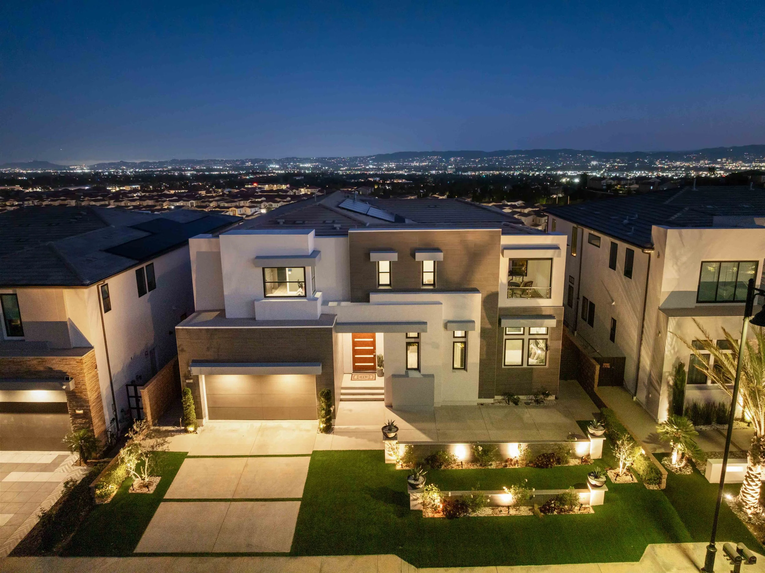 Night view of a modern two-story house with landscaped front yard, exterior lights, and neighboring houses under a dark sky with city lights visible in the distance.
