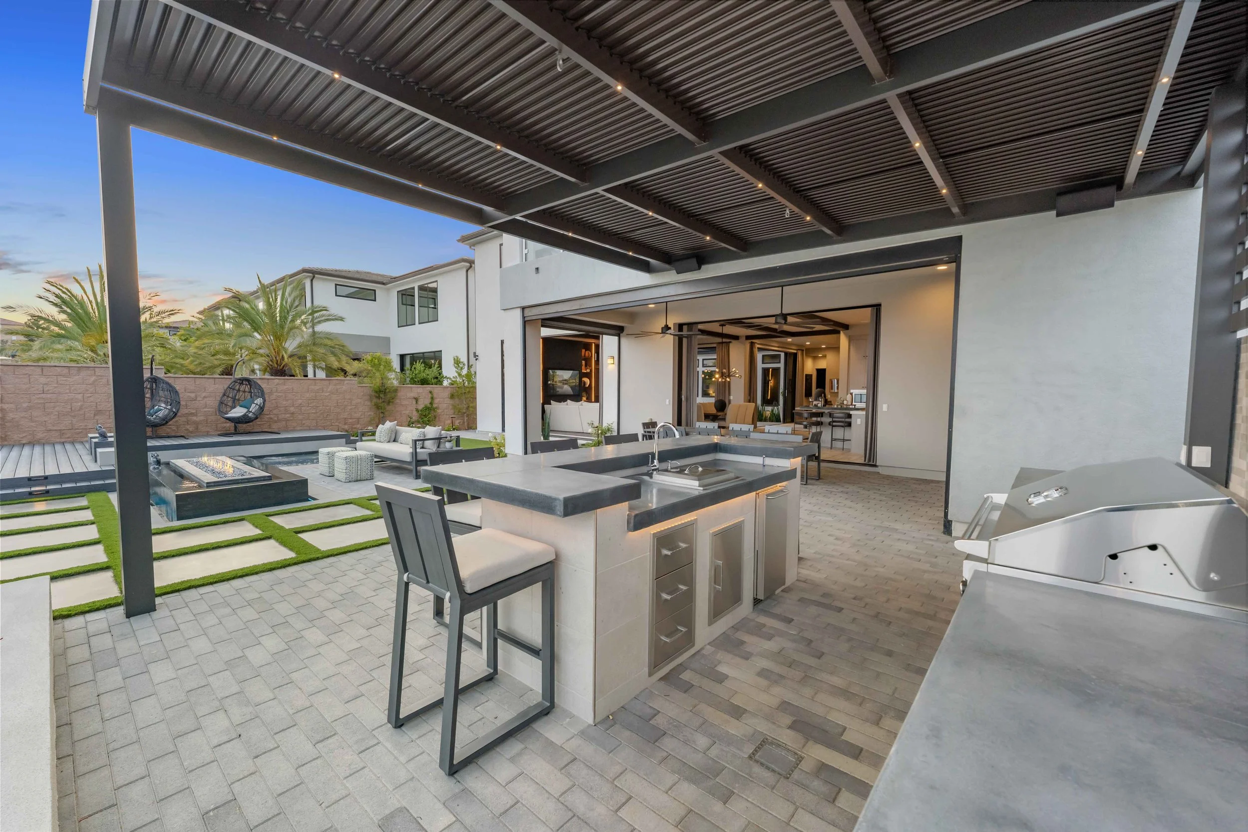 Modern outdoor kitchen and patio area with seating, fire pit, and a pool lounge with hanging basket chairs, overlooking a backyard with palm trees and a neighboring house.
