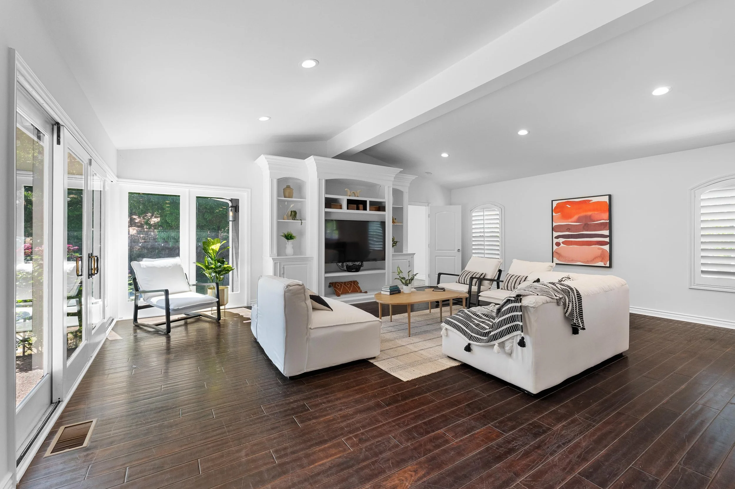 Bright living room with white sofas, dark wooden flooring, large windows with white blinds, and a wall-mounted TV surrounded by built-in shelves with decorative items. There is a modern abstract painting on the wall and a coffee table in the center.