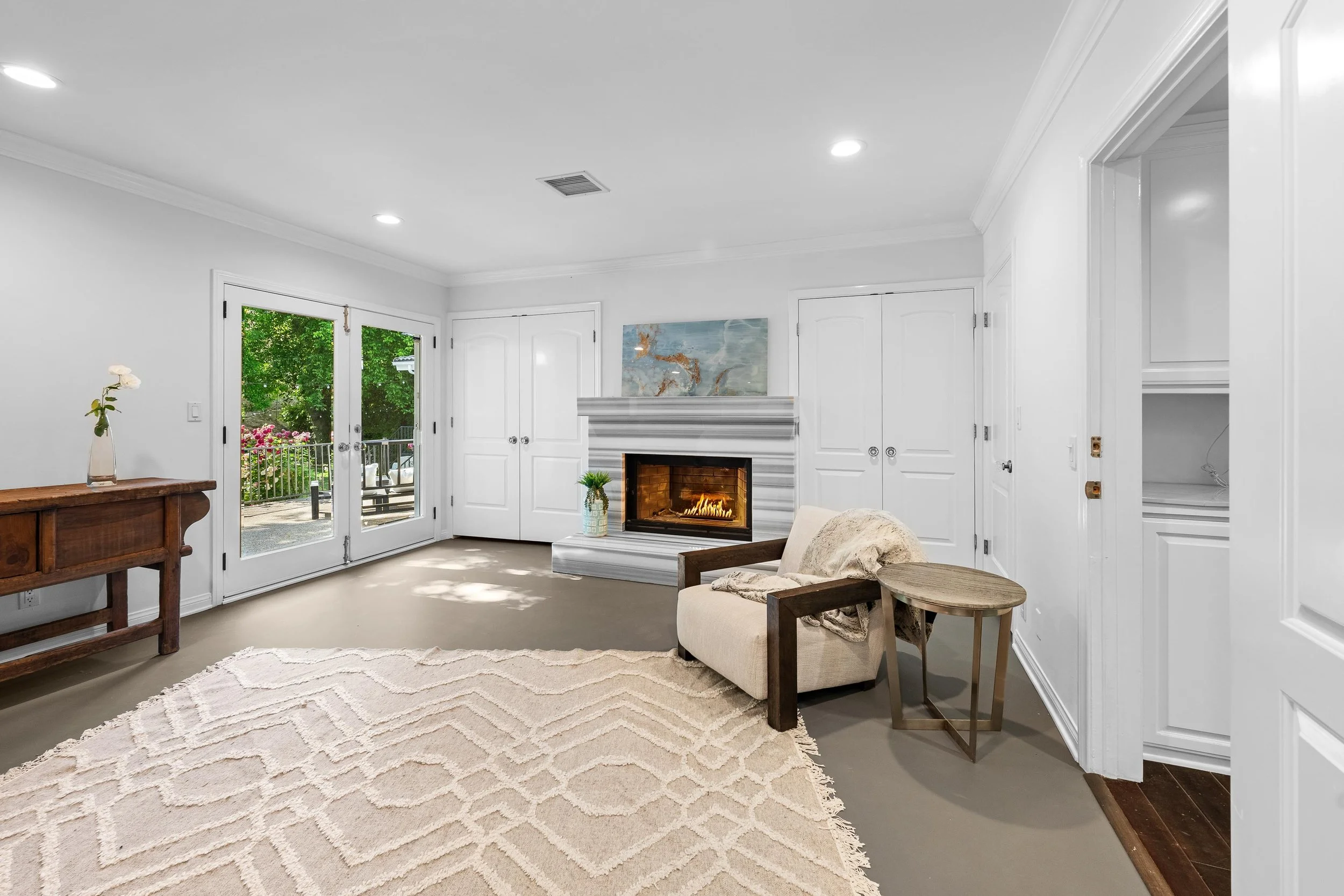 Living room with white walls, a fireplace, and a sliding glass door opening to a balcony with greenery outside. The room has a beige rug, a cream armchair with wooden arms, a small round wooden side table, and a wooden console table with a vase of fl