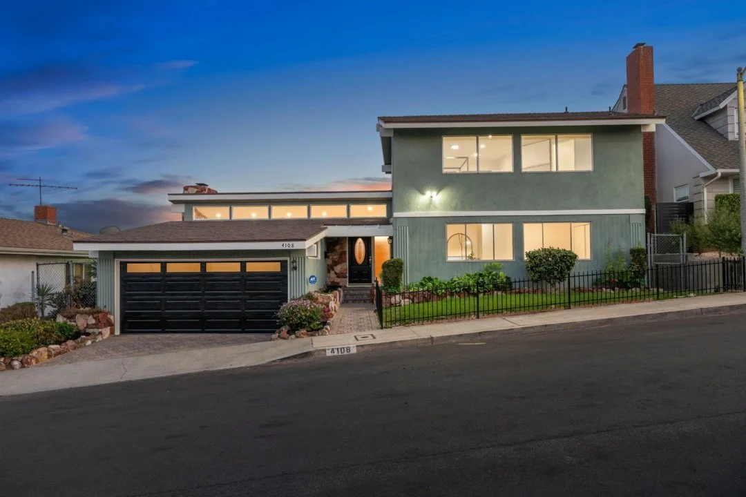 Modern two-story house with light green exterior, large windows, and a two-car garage with black doors, illuminated at dusk.