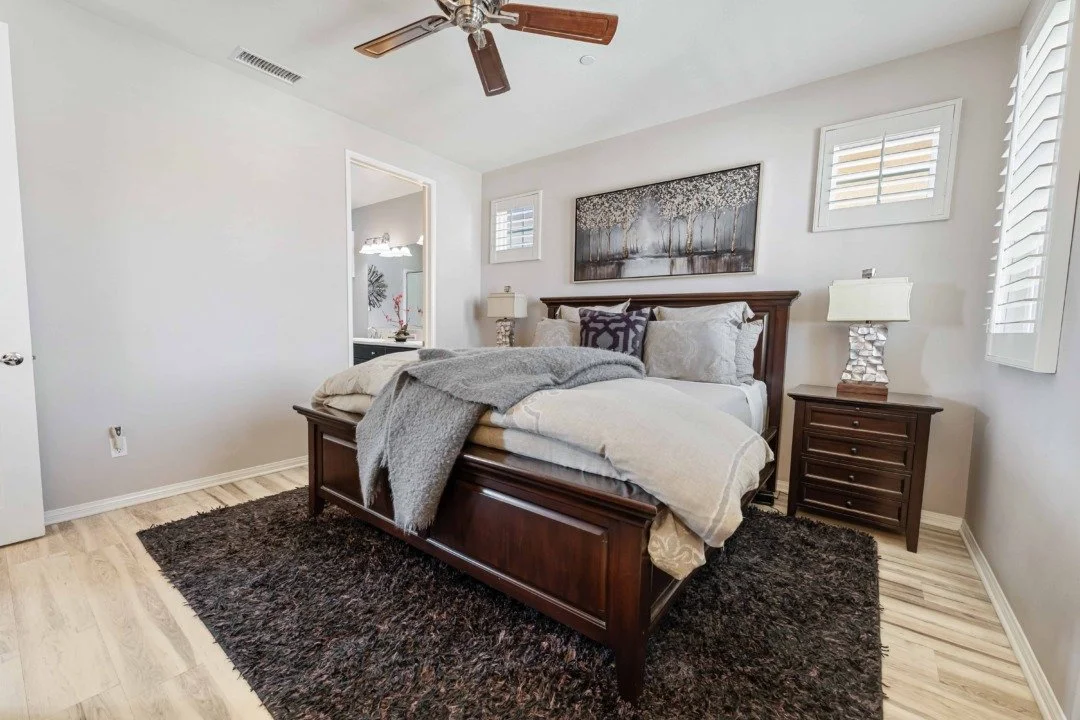 A bedroom with a wooden bed frame, a dark shaggy rug, nightstands with lamps, and wall art above the bed. There are two small windows with shutters on each side of the art, and a ceiling fan above.