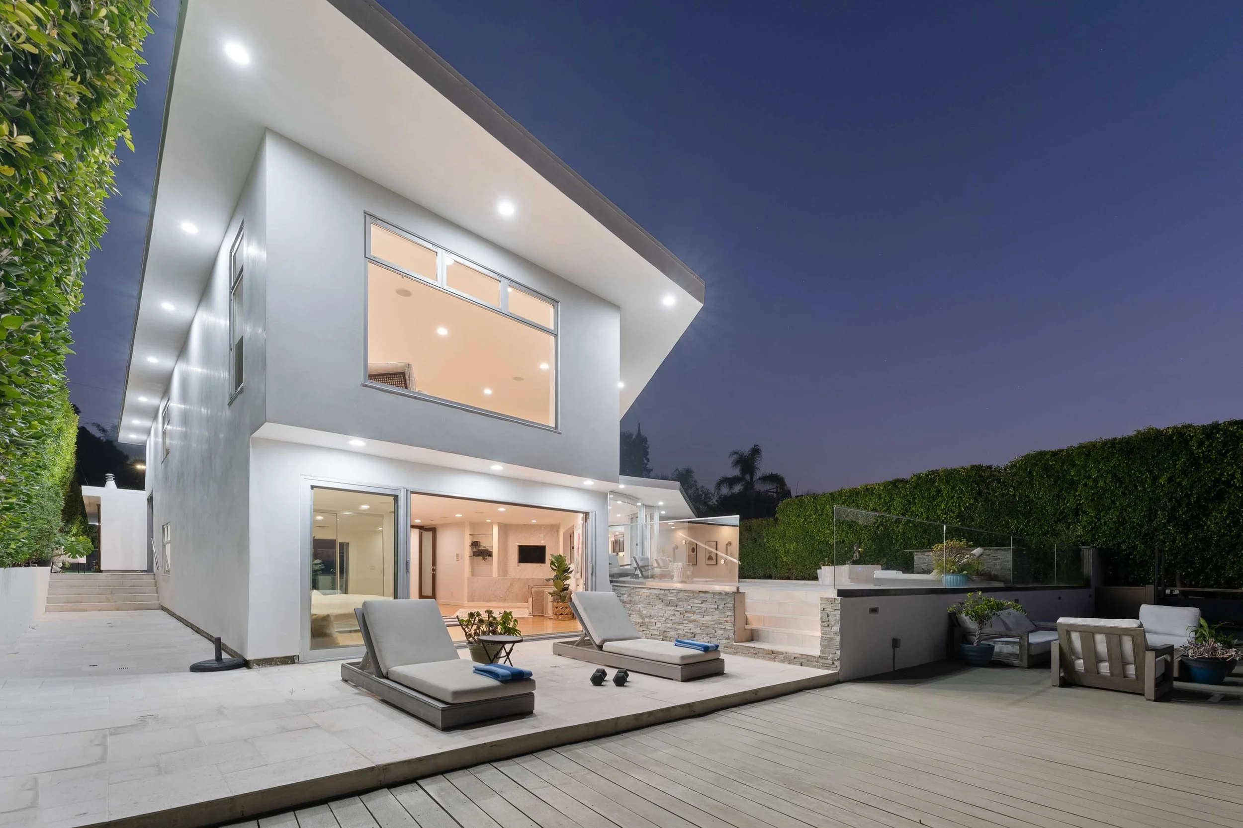 Modern two-story house at night with large glass windows, outdoor lounge chairs, potted plants, and a landscaped backyard with a wooden deck and hedges.
