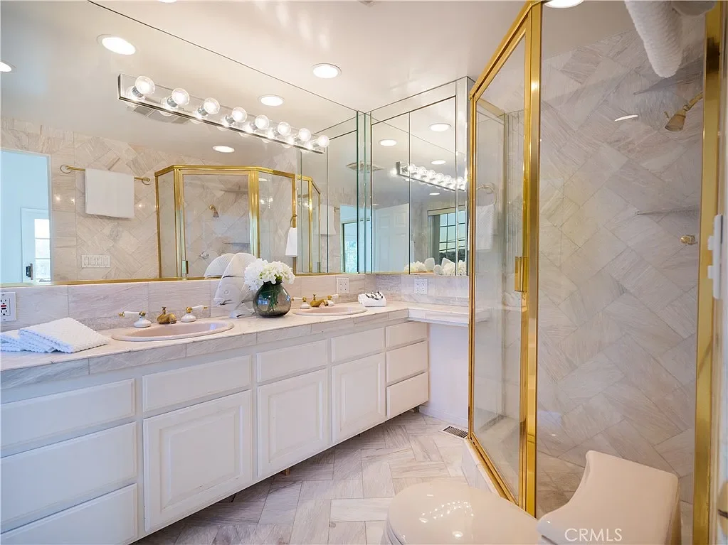 A marble bathroom with a large mirror, double sink vanity, and a glass-enclosed shower with gold accents.