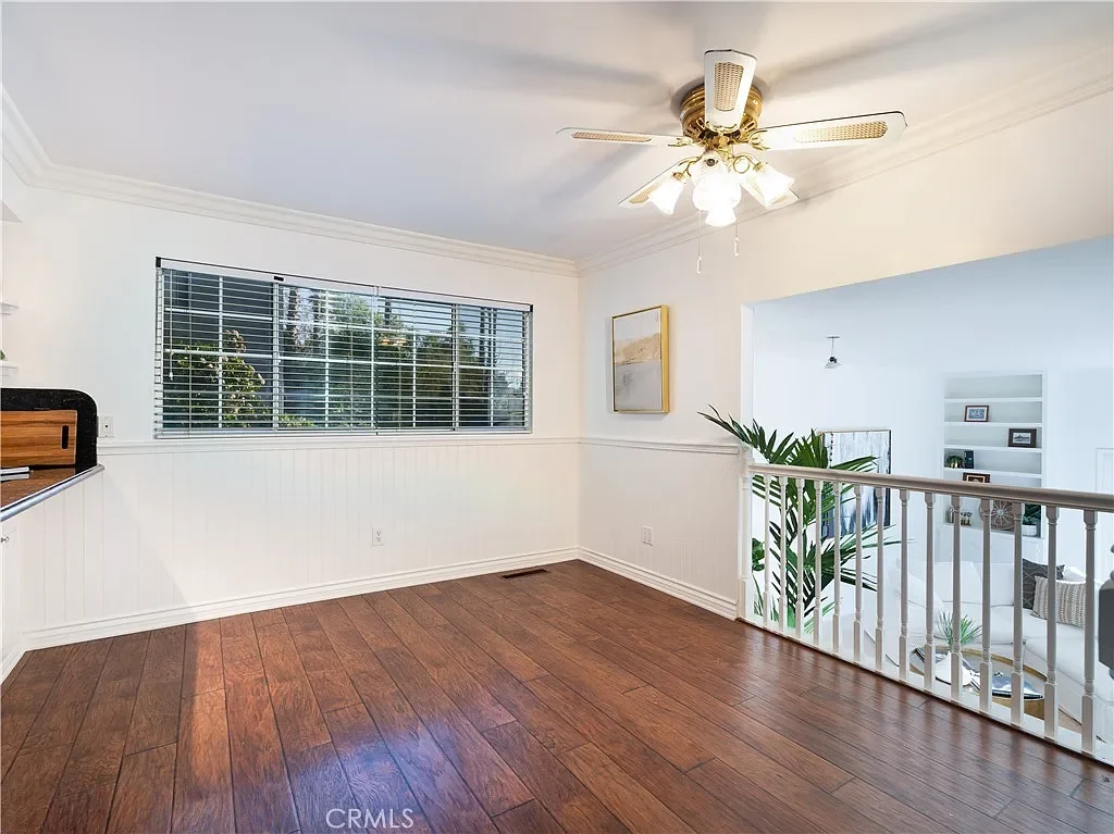 Empty room with wood flooring, white walls, a large window with blinds, a ceiling fan with lights, and a partial view of a living room with white walls, a bookshelf, and potted plants.