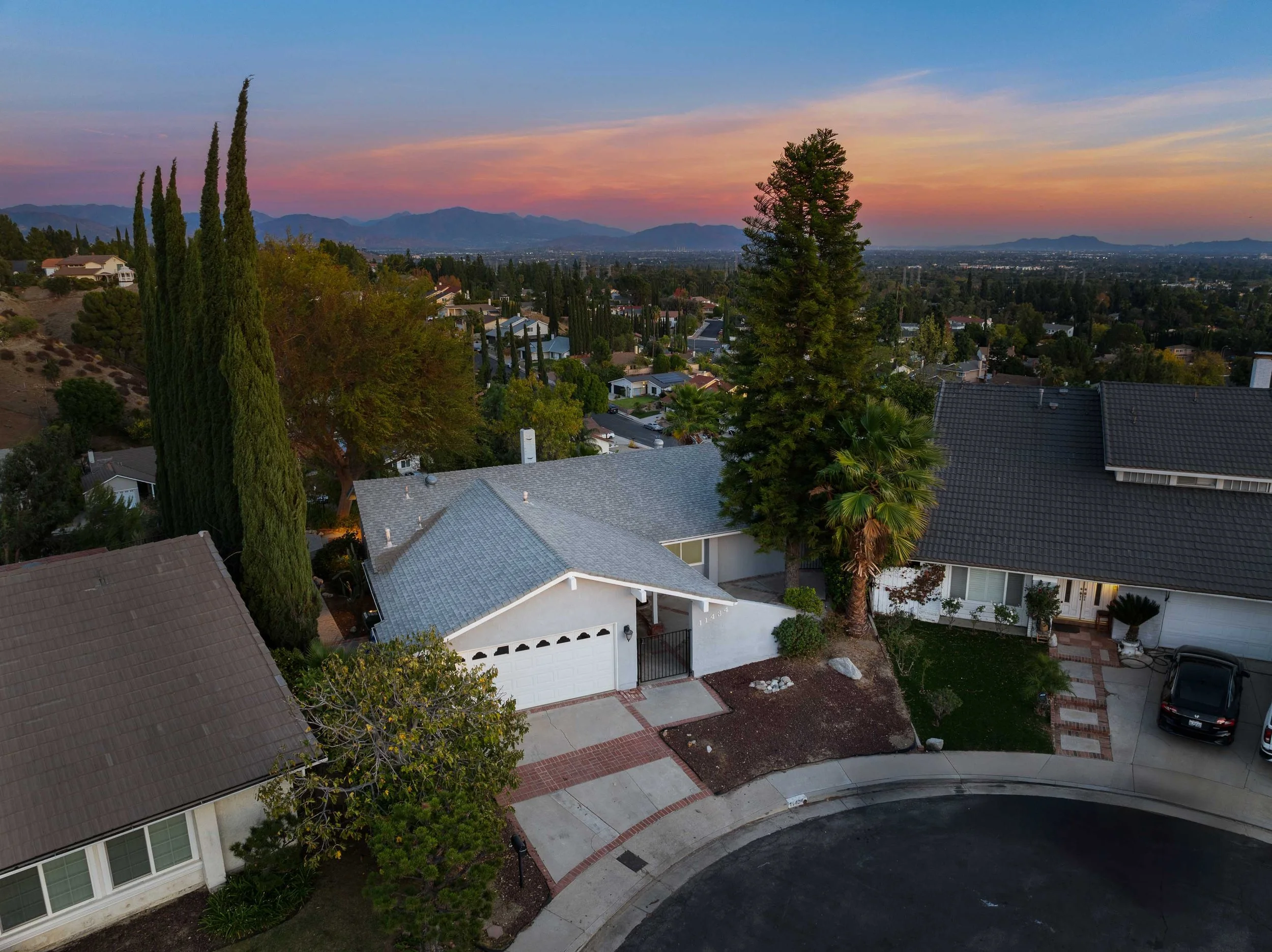 A neighborhood scene at sunset with houses, trees, and mountains in the background.