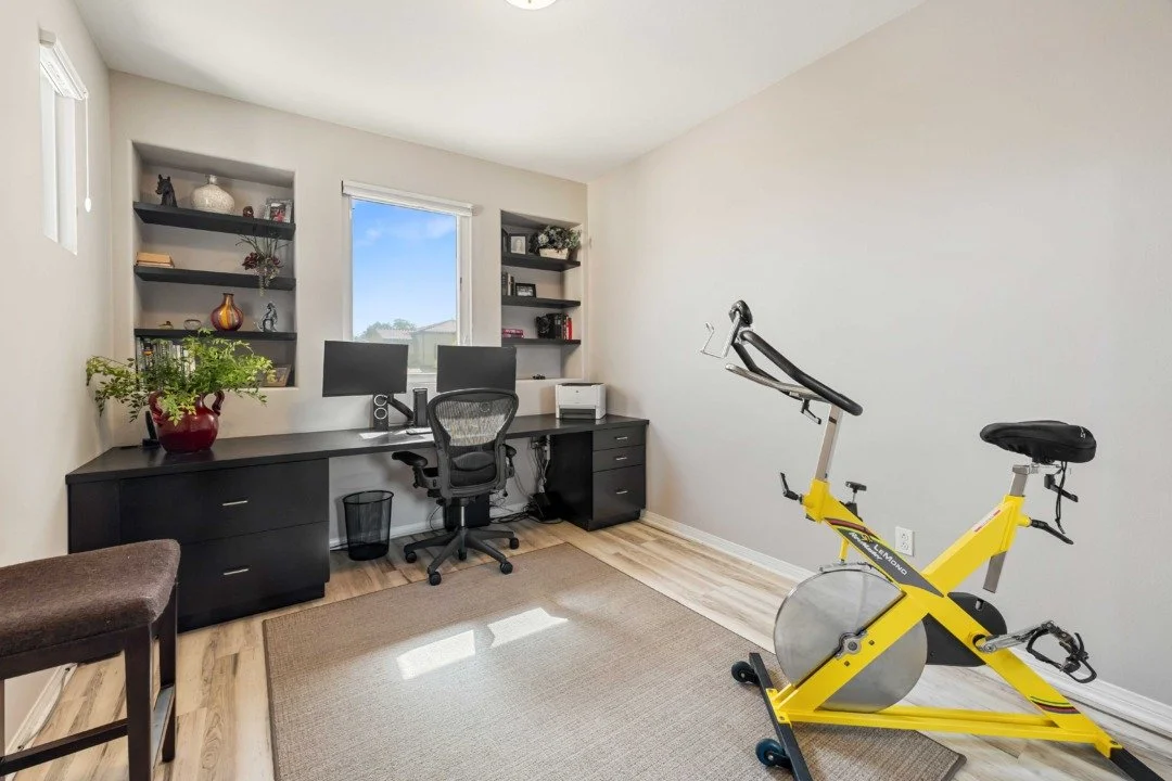 A home office with a black desk, chair, and computer monitors, facing a window. There are black shelves with decorative items on either side of the window. A yellow exercise bike is positioned against a beige wall, with a small brown rug and hardwood