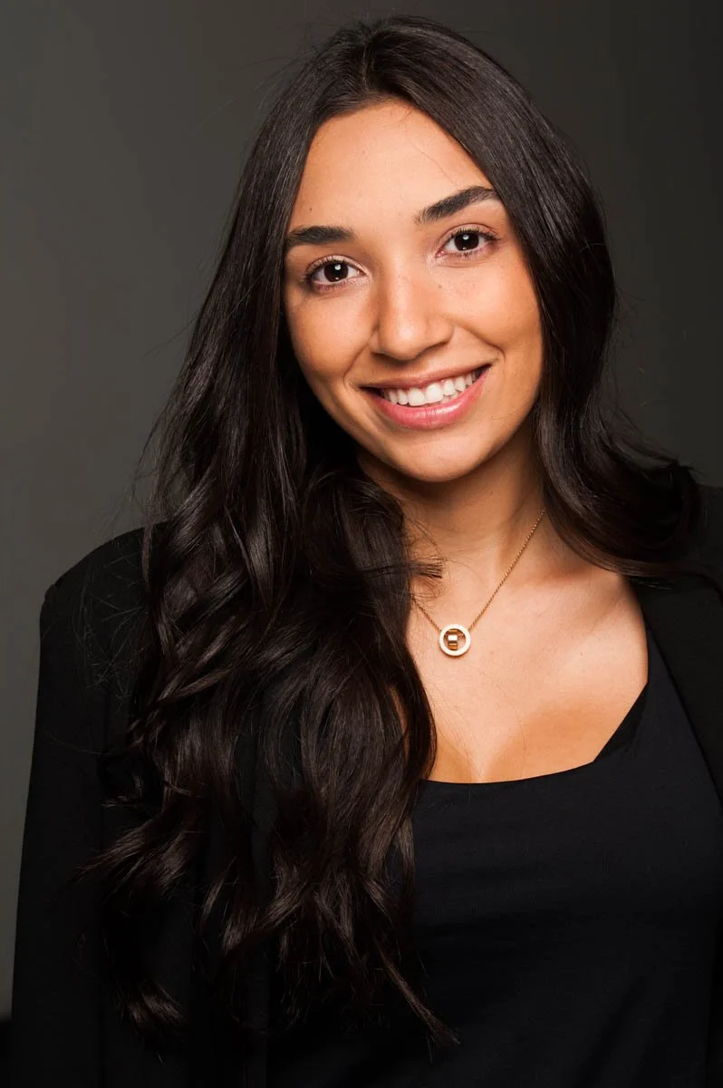 A woman with long dark hair, wearing a black top and necklace, smiling at the camera against a dark gray background.
