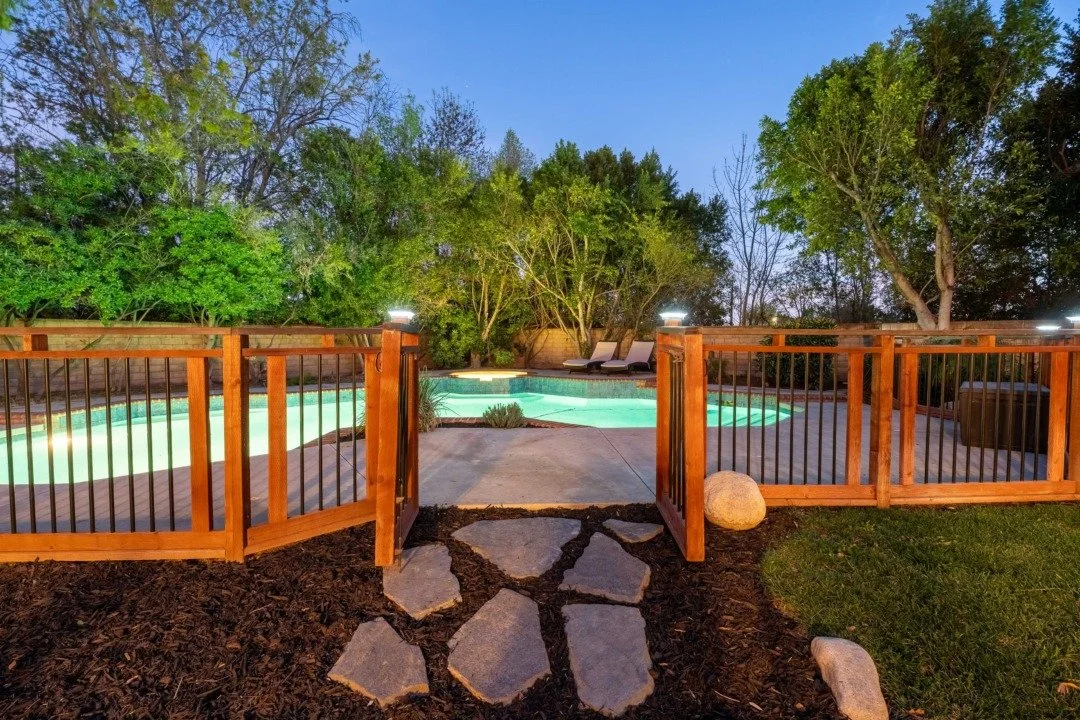 A backyard swimming pool surrounded by a wooden fence with two lounge chairs and trees in the background at dusk.