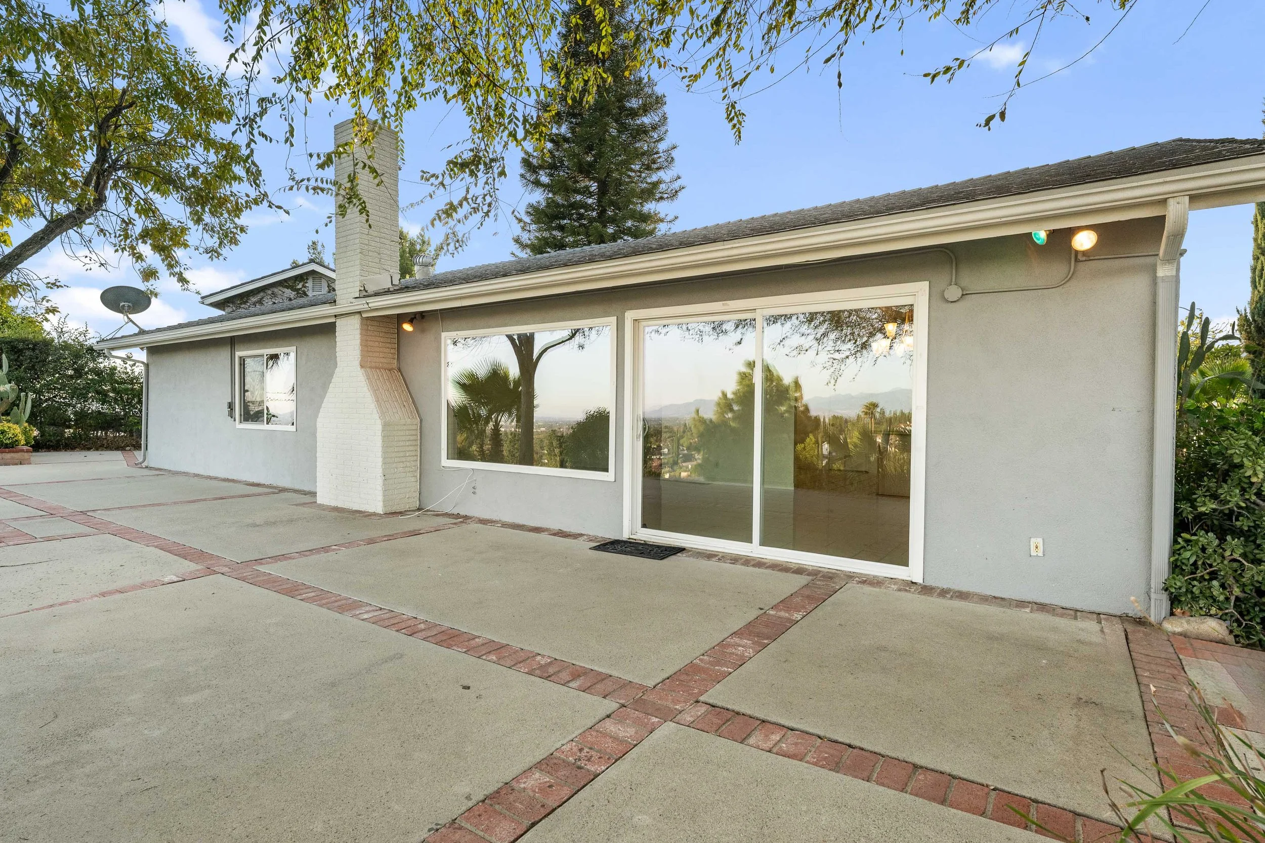 Rear view of a single-story house with a concrete patio, large sliding glass door, and a white brick chimney. Trees and mountains are visible in the background with a clear sky.