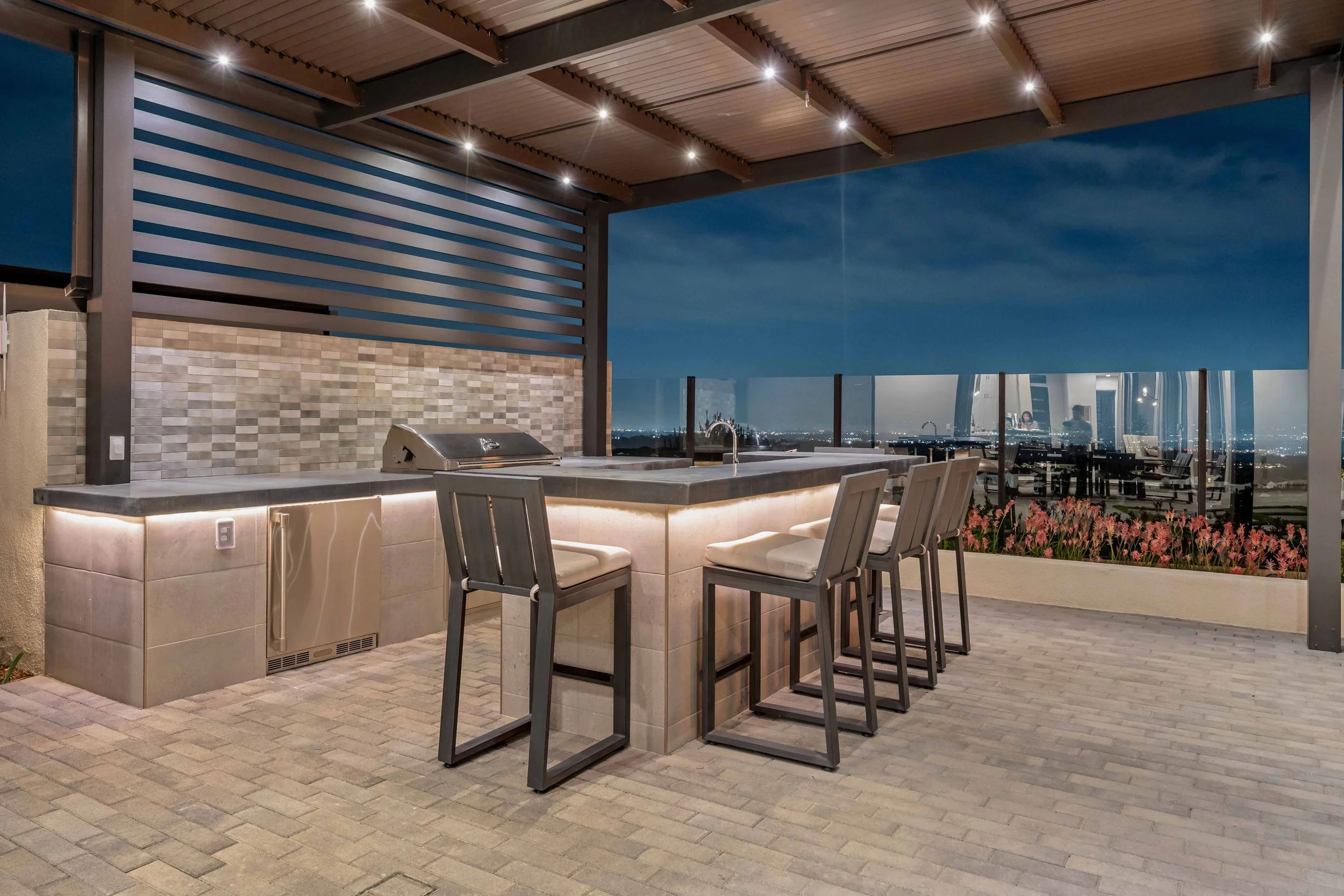 Nighttime rooftop outdoor kitchen with bar seating, built-in grill, and city skyline view through glass railing.