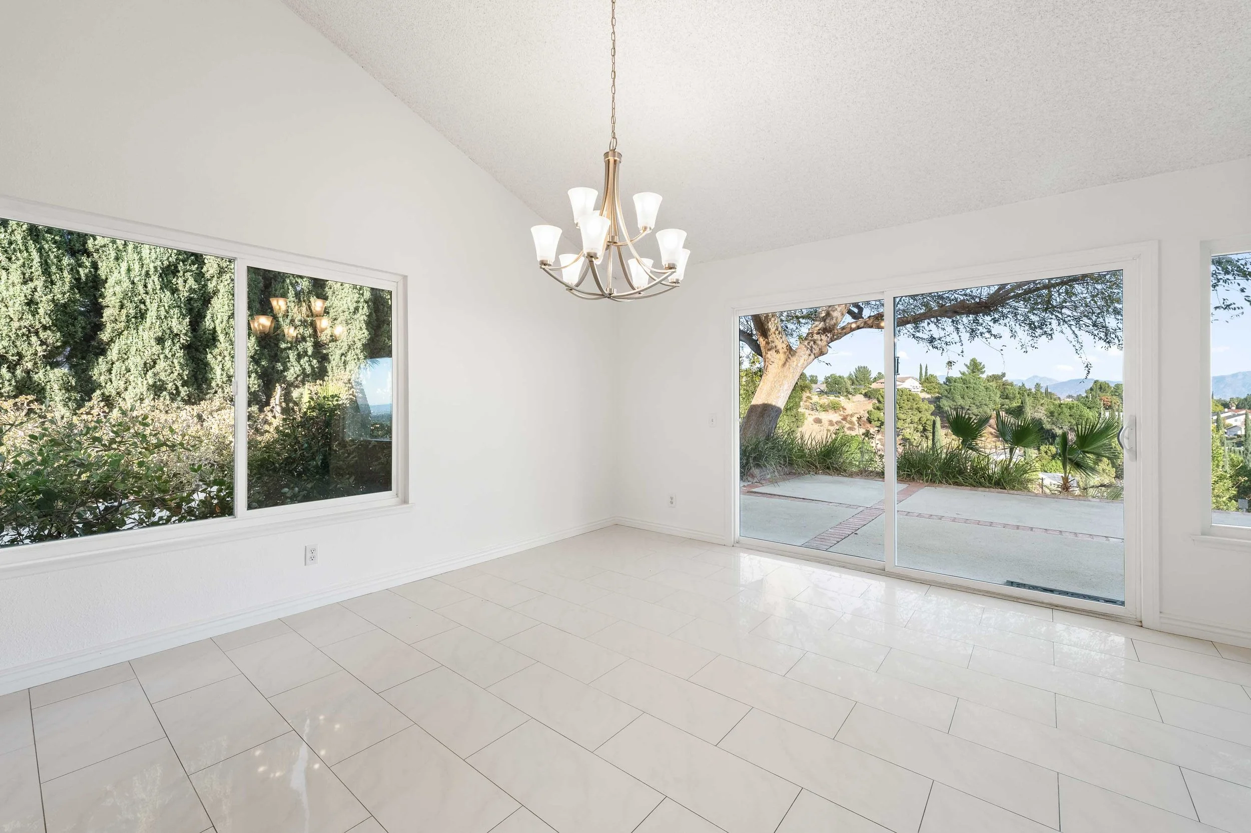 Empty bright living room with large sliding glass doors and windows, white walls, tiled floor, view of outdoor patio and greenery, and a chandelier hanging from the ceiling.
