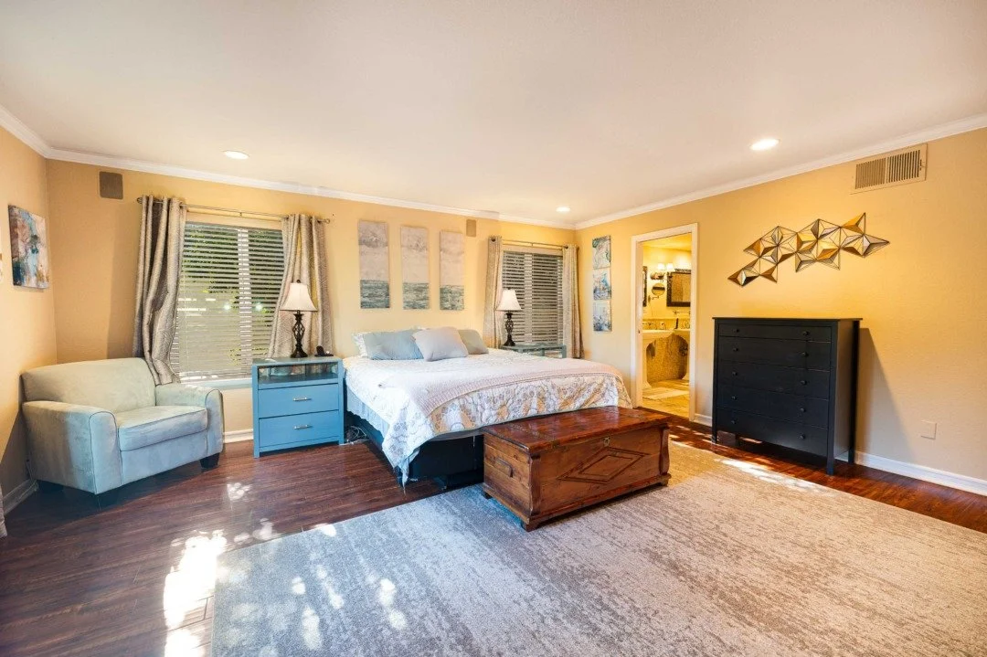 A bedroom with a large bed, two nightstands with lamps, a light green armchair, a blue chest of drawers, and a wooden trunk at the foot of the bed. There are three paintings above the bed, windows with curtains on either side, and a doorway leading t