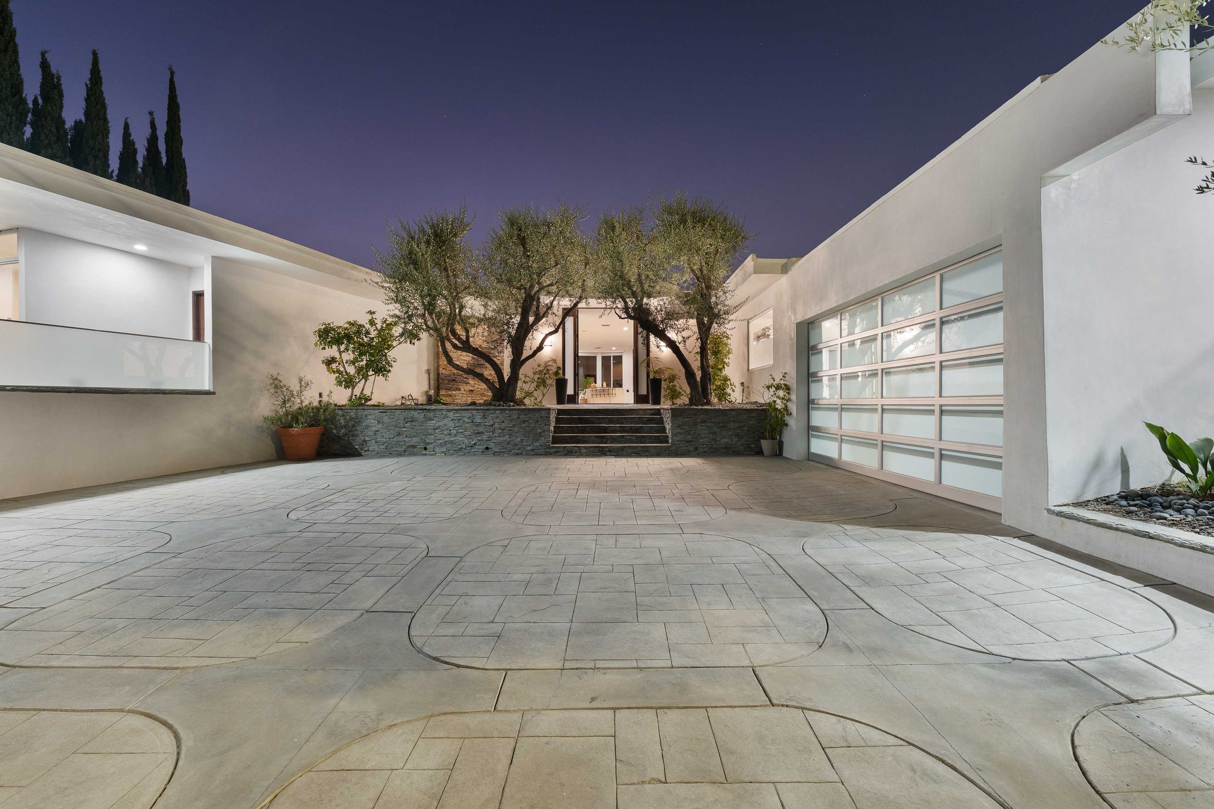 Modern white house with large glass garage door, outdoor courtyard, and trees illuminated at night.
