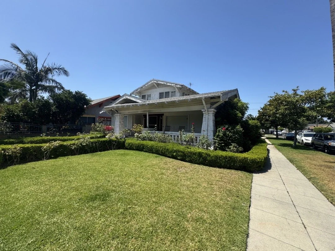 A white, two-story house with a front porch, surrounded by a well-maintained lawn, bushes, and trees, with a sidewalk and parked cars along the street.