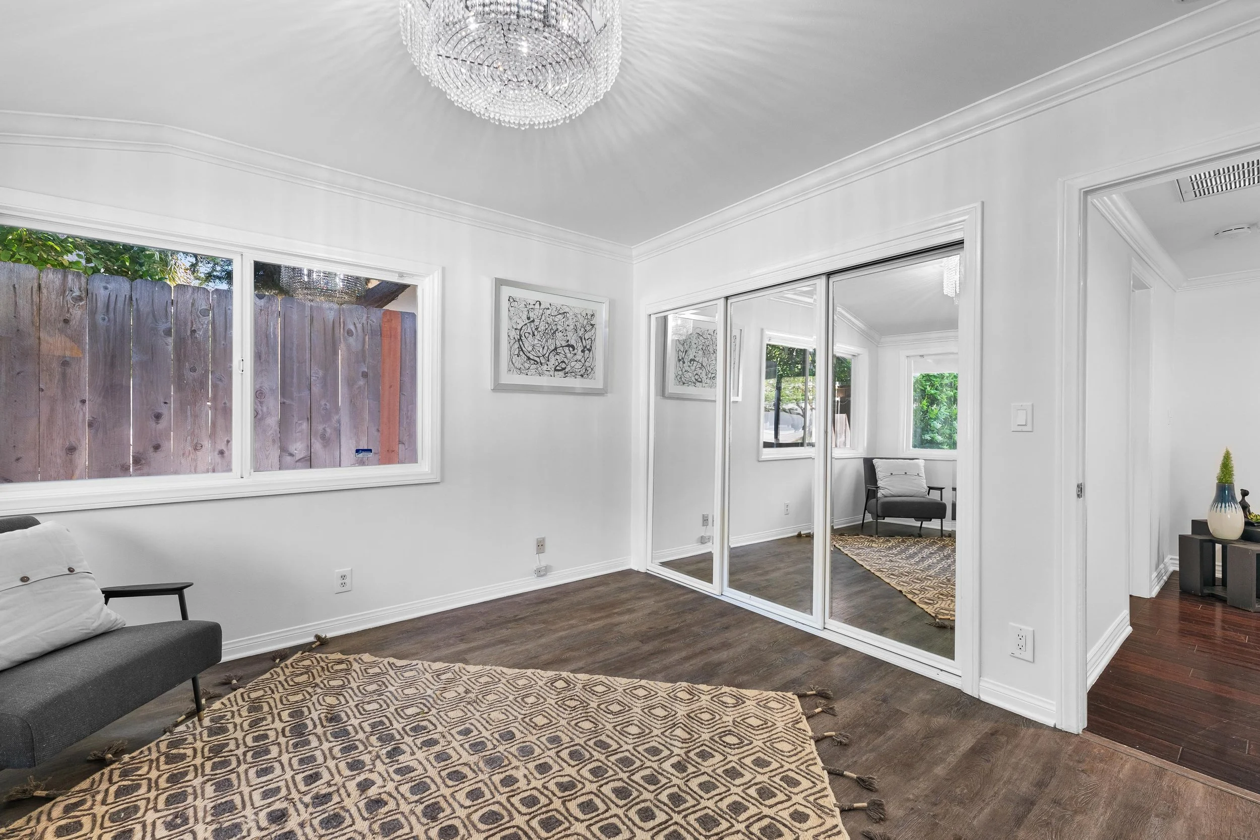 Empty living room with white walls, large window, and mirrored sliding closet doors. Contains a small gray sofa with white pillow, decorative rug, and minimal artwork. Adjacent room visible through doorway and mirror.