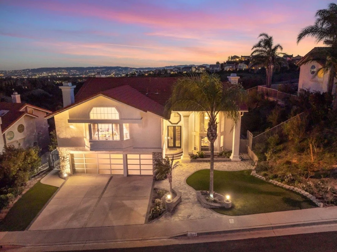 A two-story house with a red-tiled roof and a beige exterior, illuminated at dusk, with a driveway, palm trees, and landscaped yard.