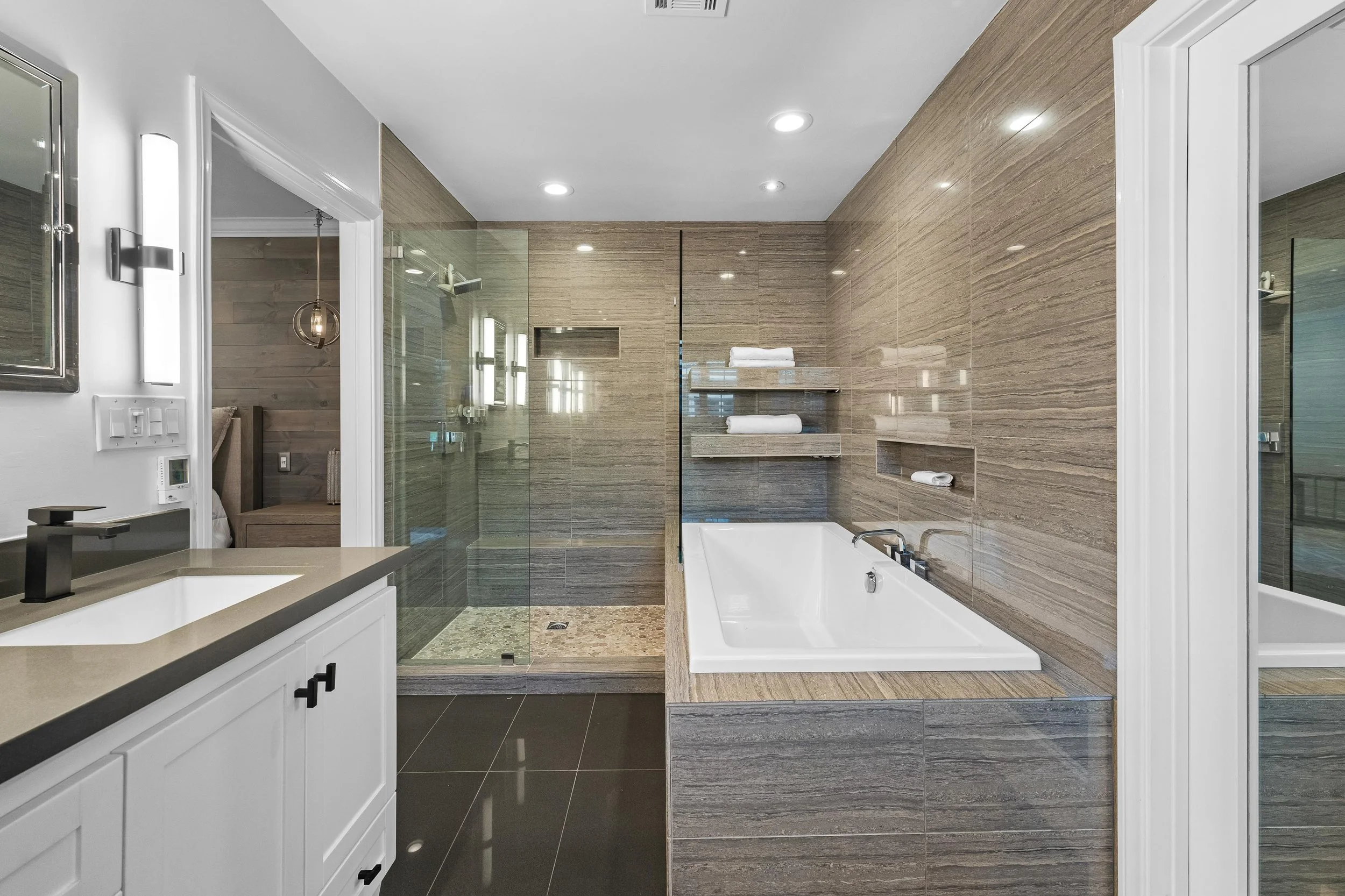 Modern bathroom with a white vanity, black faucet, beige countertop, and a large bathtub with a wood-like surround. Behind the tub, there are built-in shelves with towels and a wide glass shower with stone tiles and a glass door.