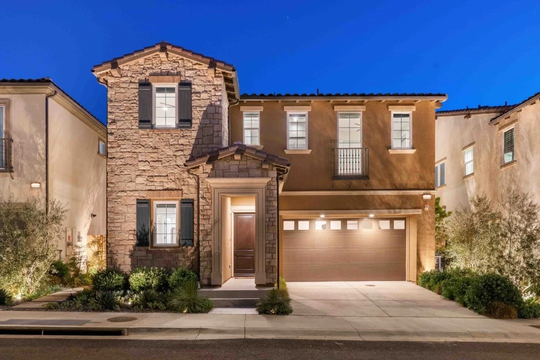 A two-story house with stone and stucco exterior, illuminated at night, featuring a front door, garage, and several windows with shutters.
