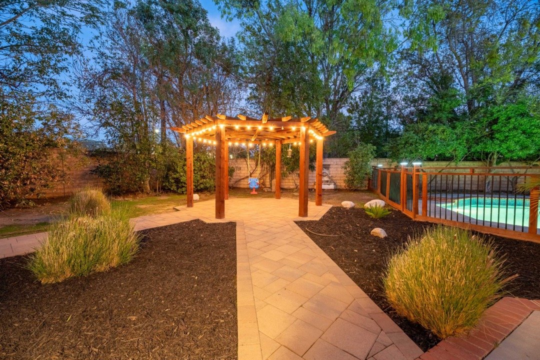 A backyard patio with a lit pergola, a small blue swing hanging underneath, a stone pathway, planted bushes, and a fenced-in pool area in the evening.