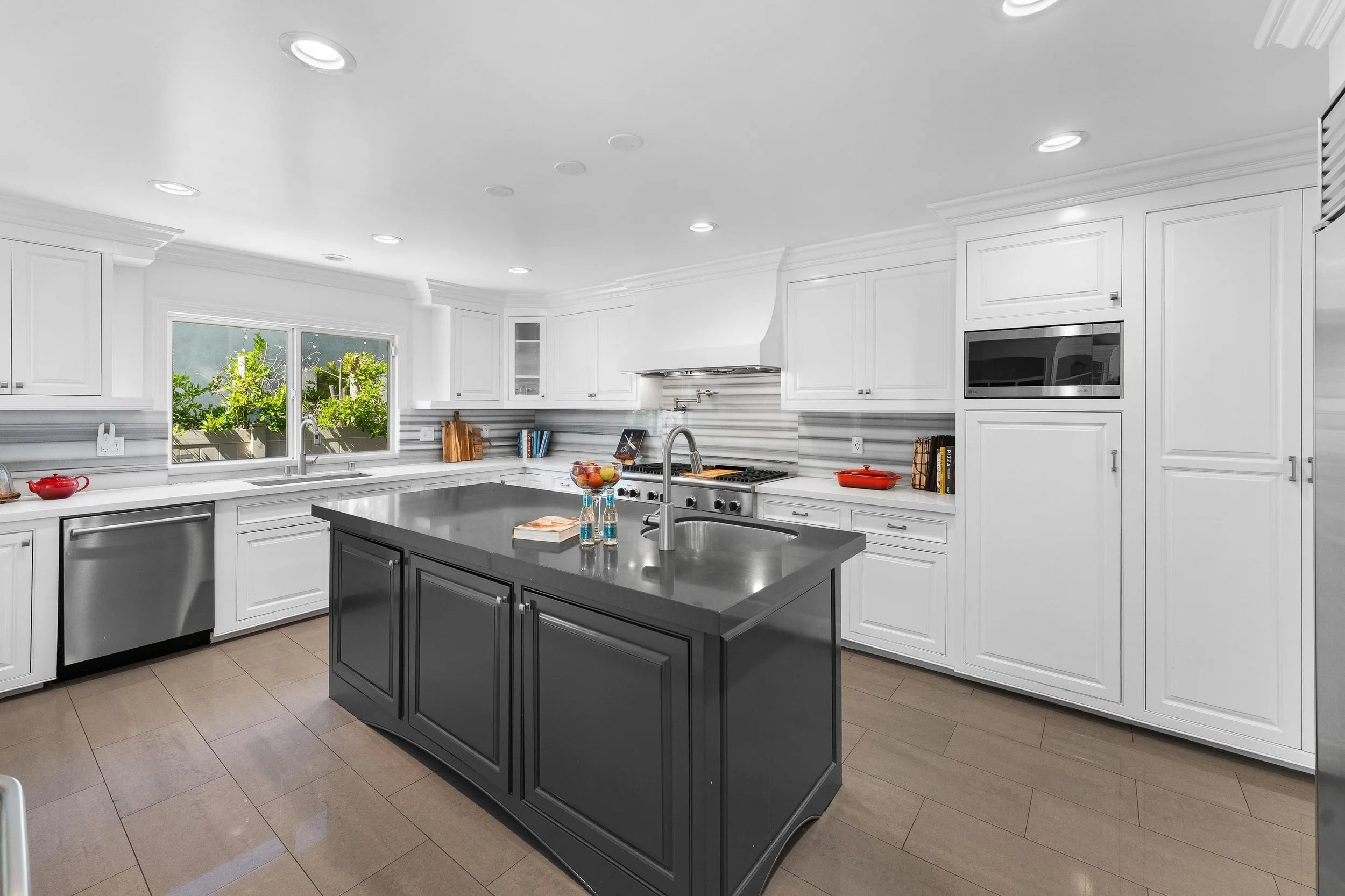 Modern kitchen with white cabinets, a black island, stainless steel appliances, and a window showing green foliage outside.