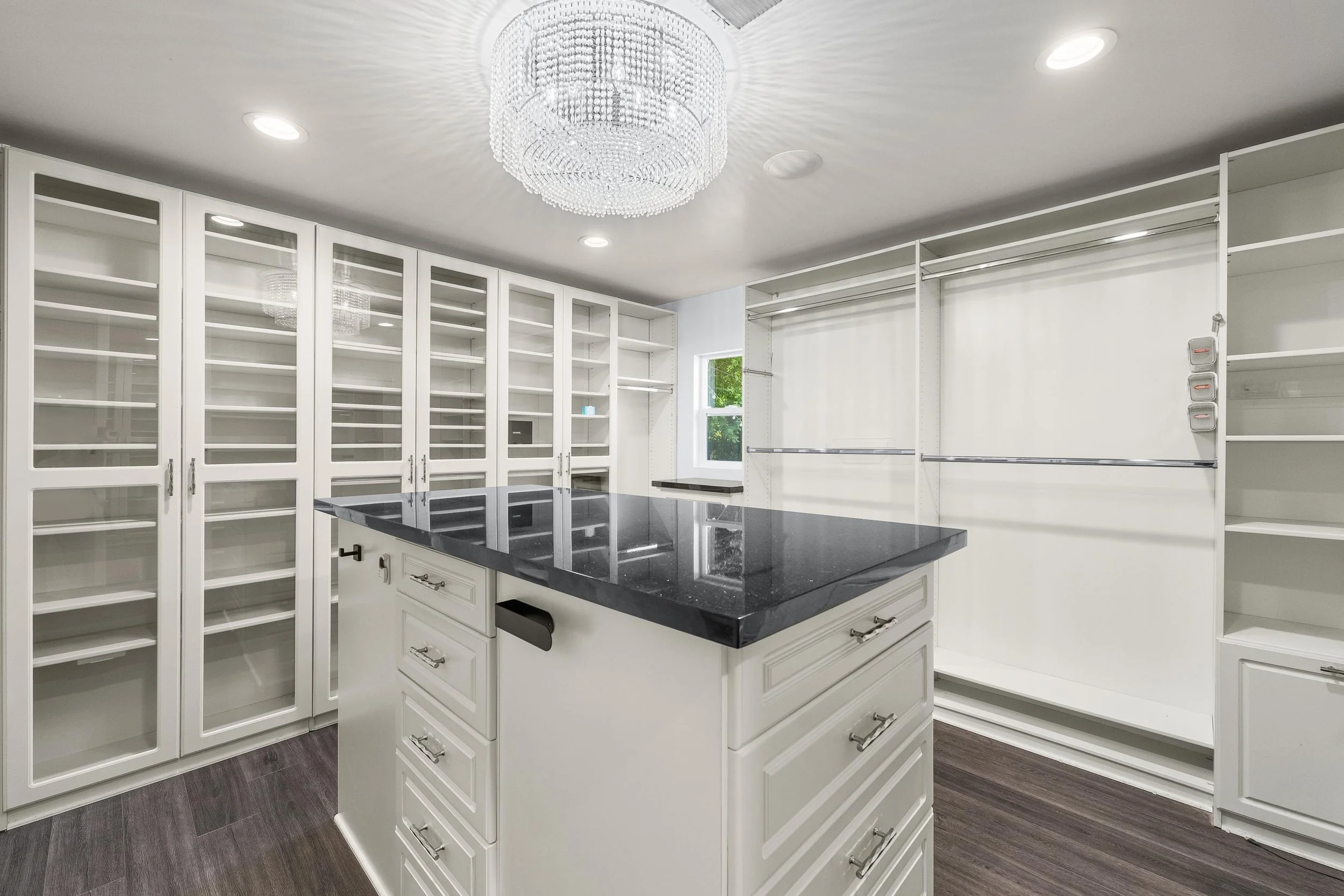 A spacious, empty walk-in closet with white cabinets and shelves, a black countertop on a central island, and a small window.