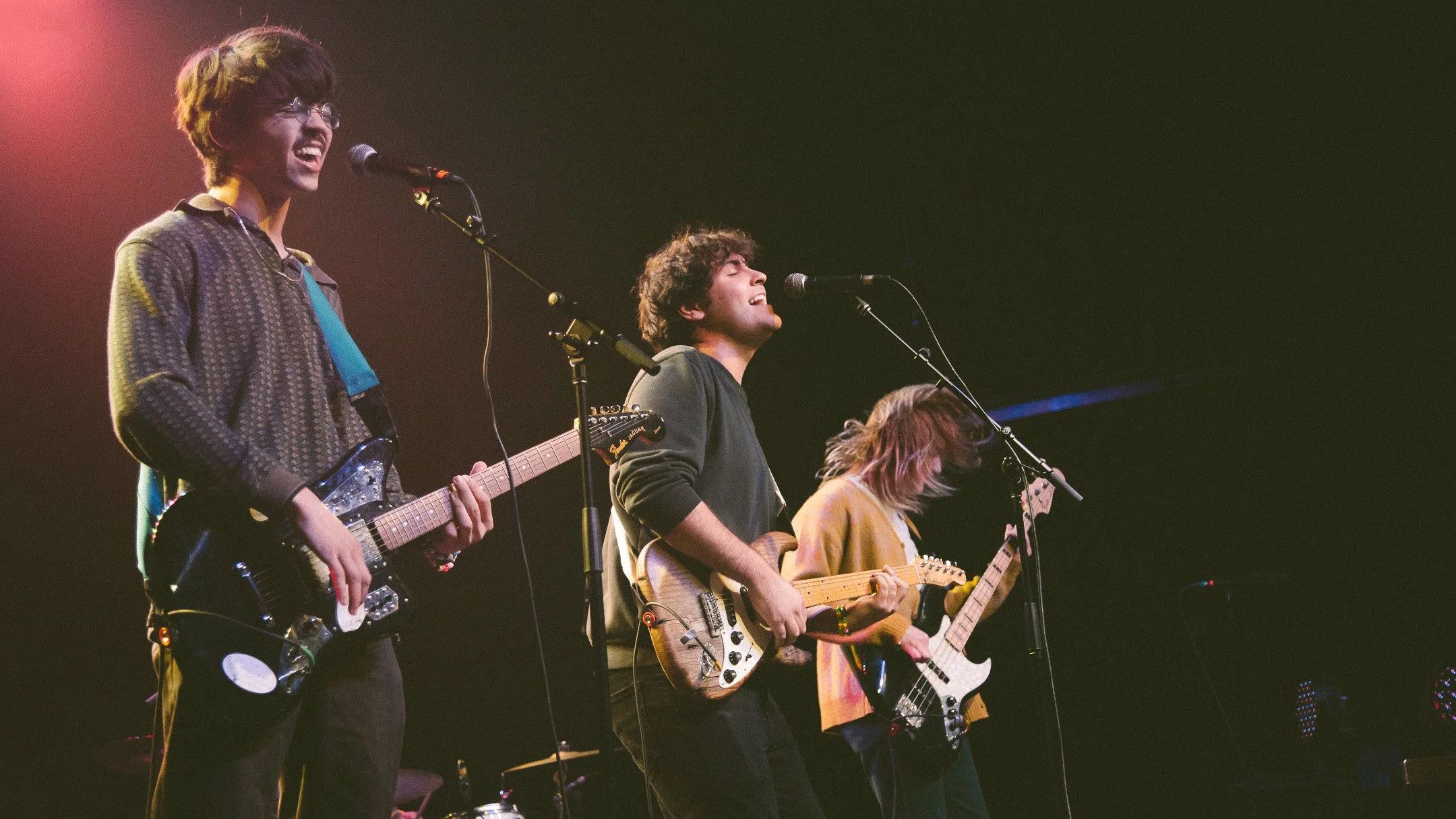 Three young musicians performing on stage with guitars and microphones, singing passionately under stage lights.