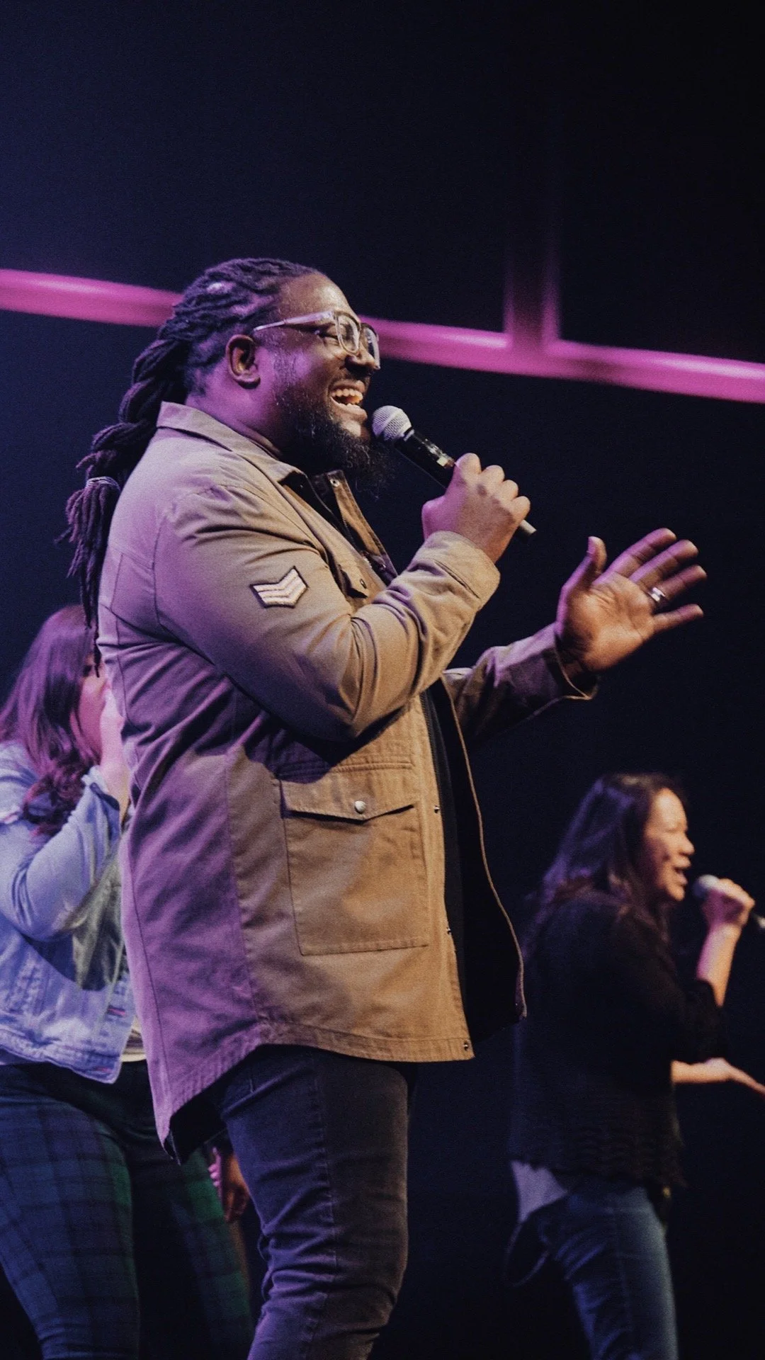 A man with dreadlocks and glasses singing into a microphone, with two women in the background also singing, on stage under purple lighting.
